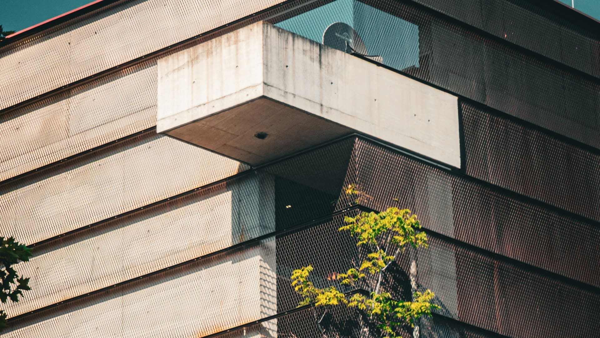 Modern building with a geometric concrete balcony jutting out from metal mesh walls, partially shaded, with green tree branches in the lower right foreground.