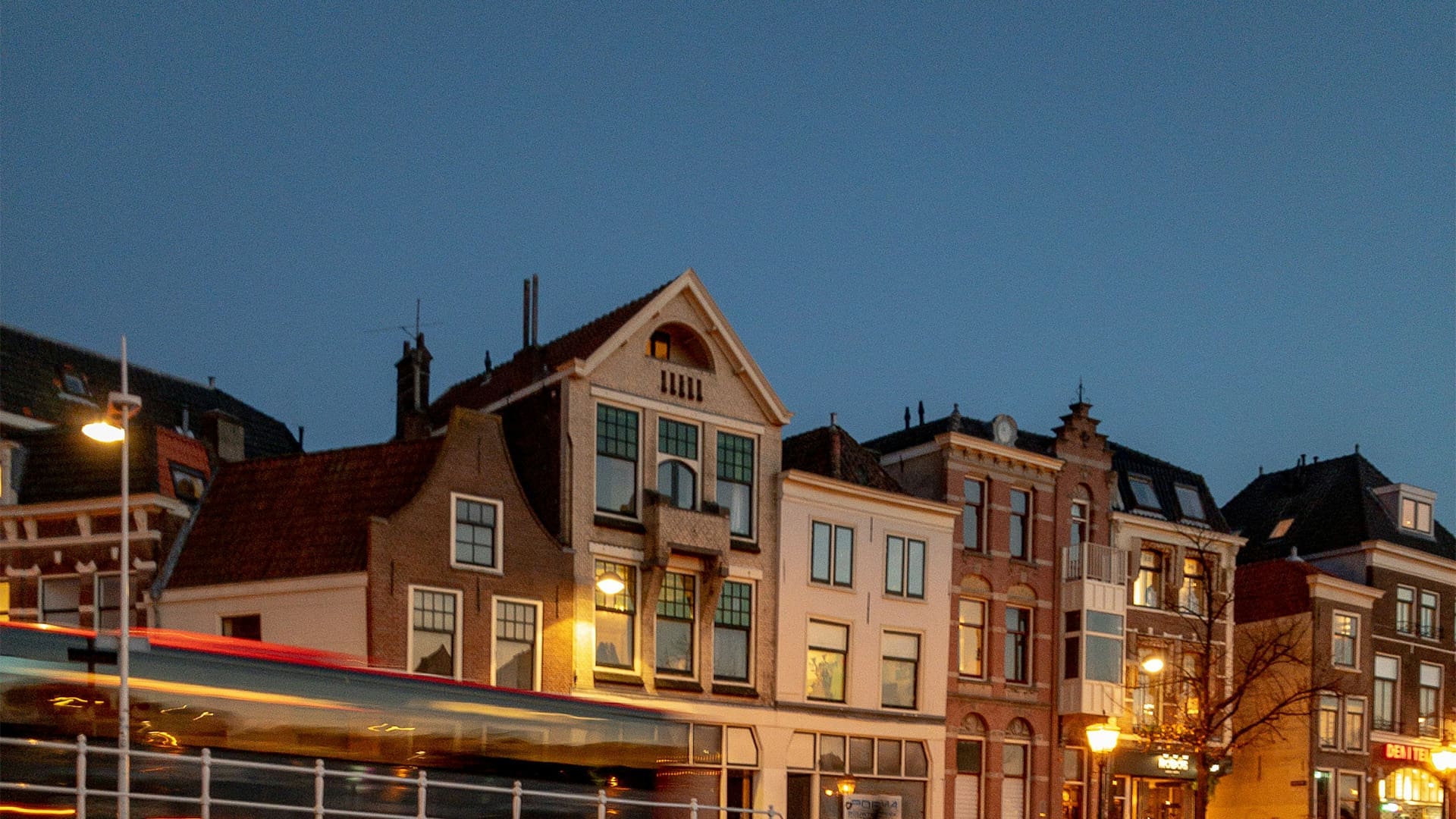A row of traditional European buildings at dusk, with warm lights glowing in windows. In the foreground, a tram or bus passes by, appearing blurred due to motion. The sky is clear and deep blue.