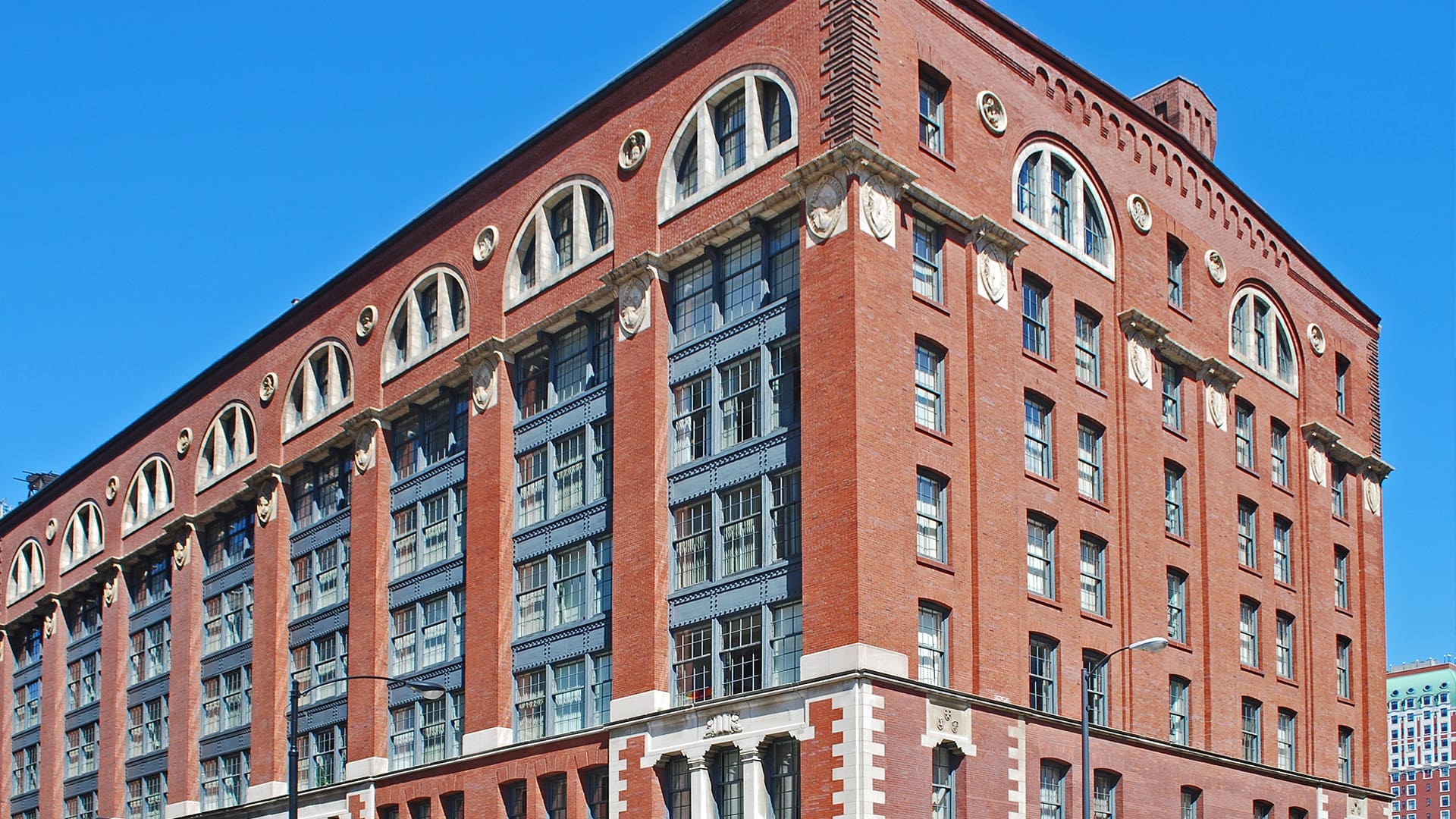 A red-brick multi-story building with arched windows, decorative stonework, and large gray-framed windows, set against a clear blue sky.