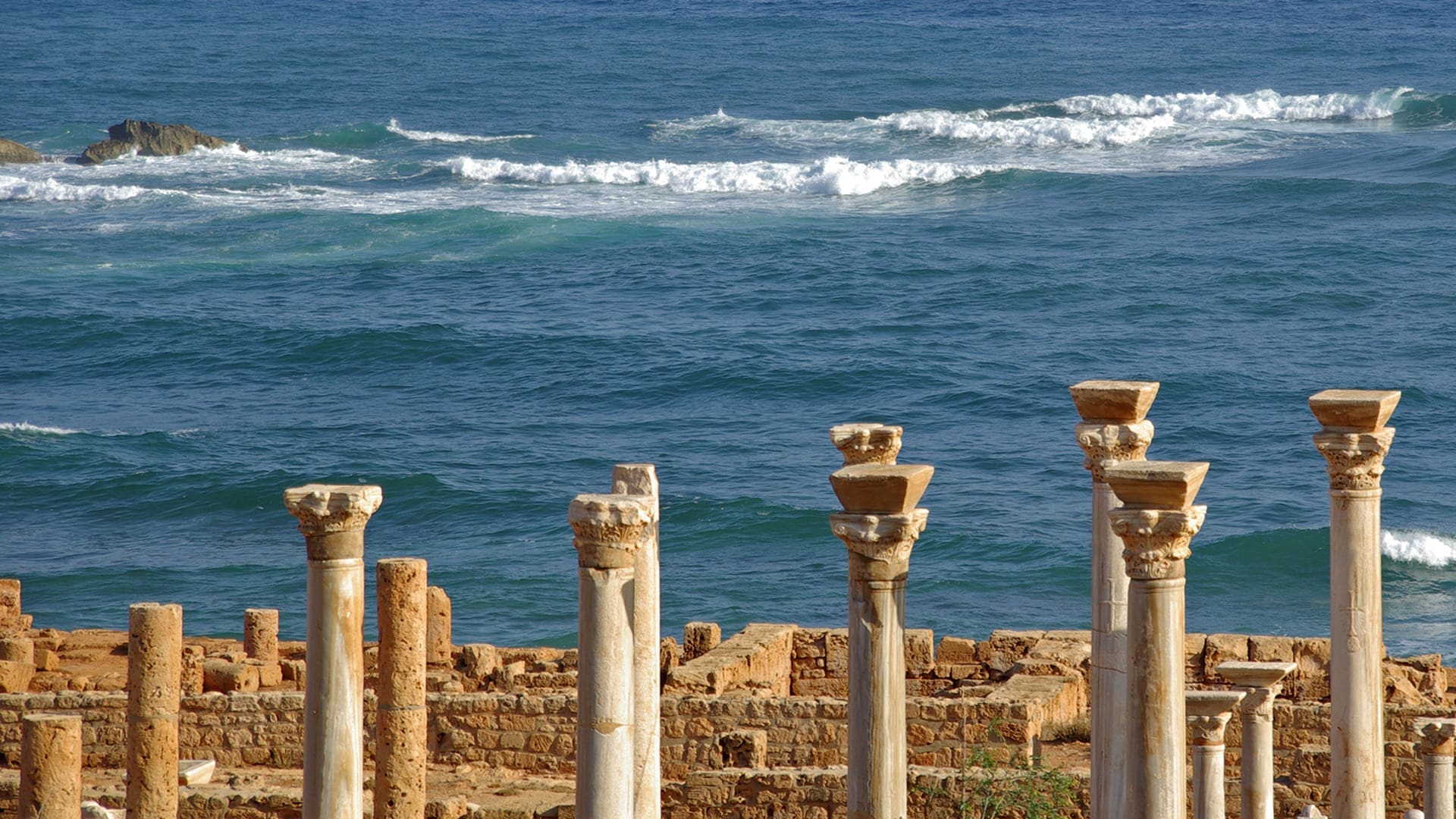 Ancient stone columns stand in front of ruins by the sea, with blue waves and white foam in the background under a clear sky.