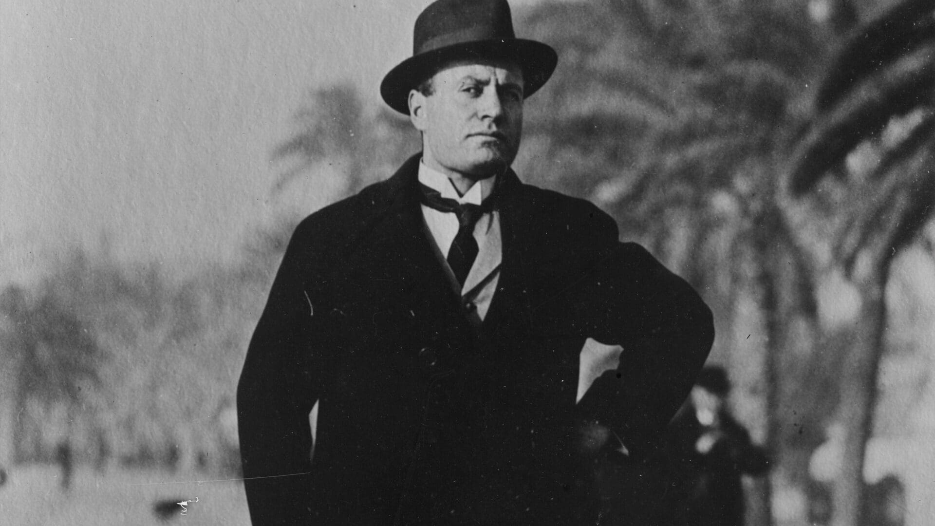 A man in a dark overcoat and hat stands outdoors with his hand on his hip. Trees and blurred figures are visible in the background. The image is black and white.