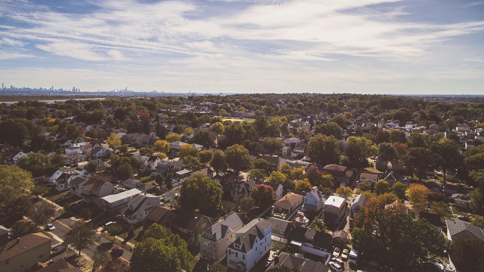 Aerial view of a suburban neighborhood with many houses surrounded by trees under a partly cloudy sky, with a city skyline visible in the distant background.