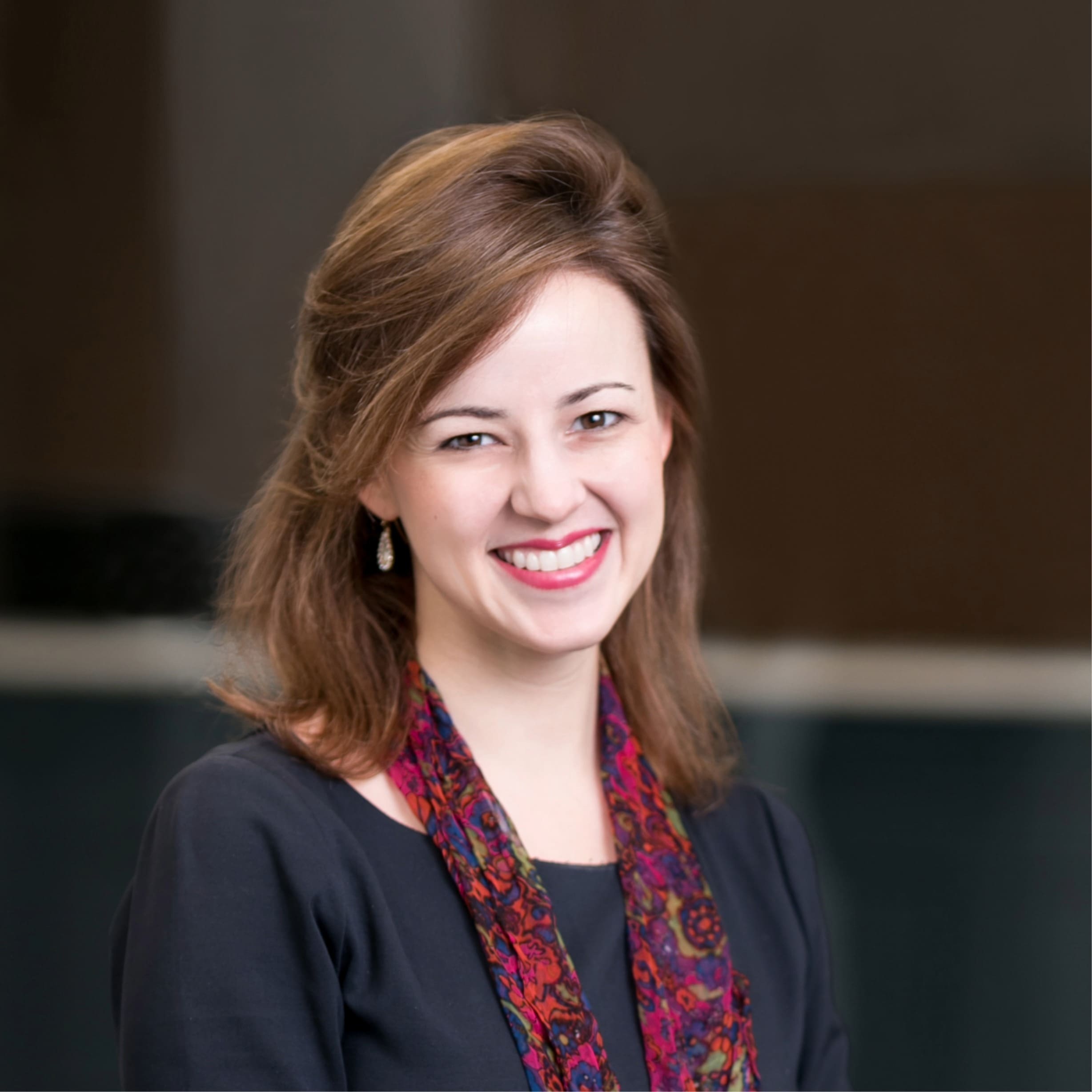 A woman with light brown hair and a patterned scarf smiles at the camera. She is wearing a dark top and small earrings, with a blurred dark background behind her.