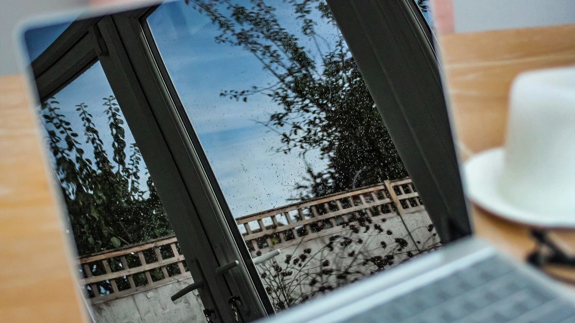 A laptop screen reflects a glass door, trees, and a cloudy sky. Nearby, a white cup and saucer sit on a wooden table. The outdoor scene is visible through the reflection on the laptop display.