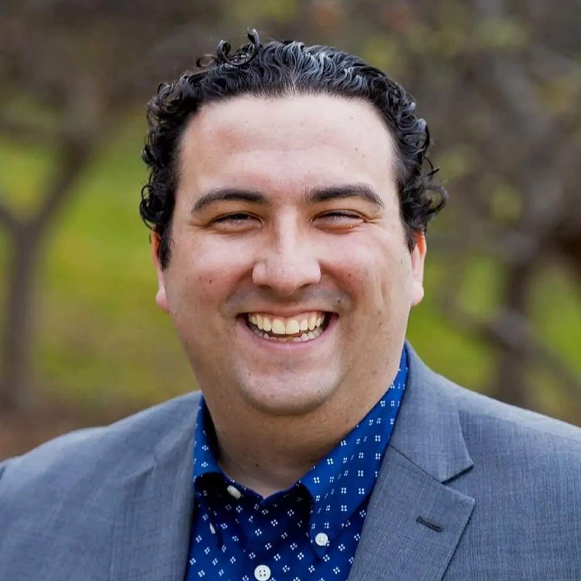 A man with curly dark hair wearing a gray suit jacket and a blue patterned shirt is smiling broadly outdoors, with a blurred background of greenery and trees.