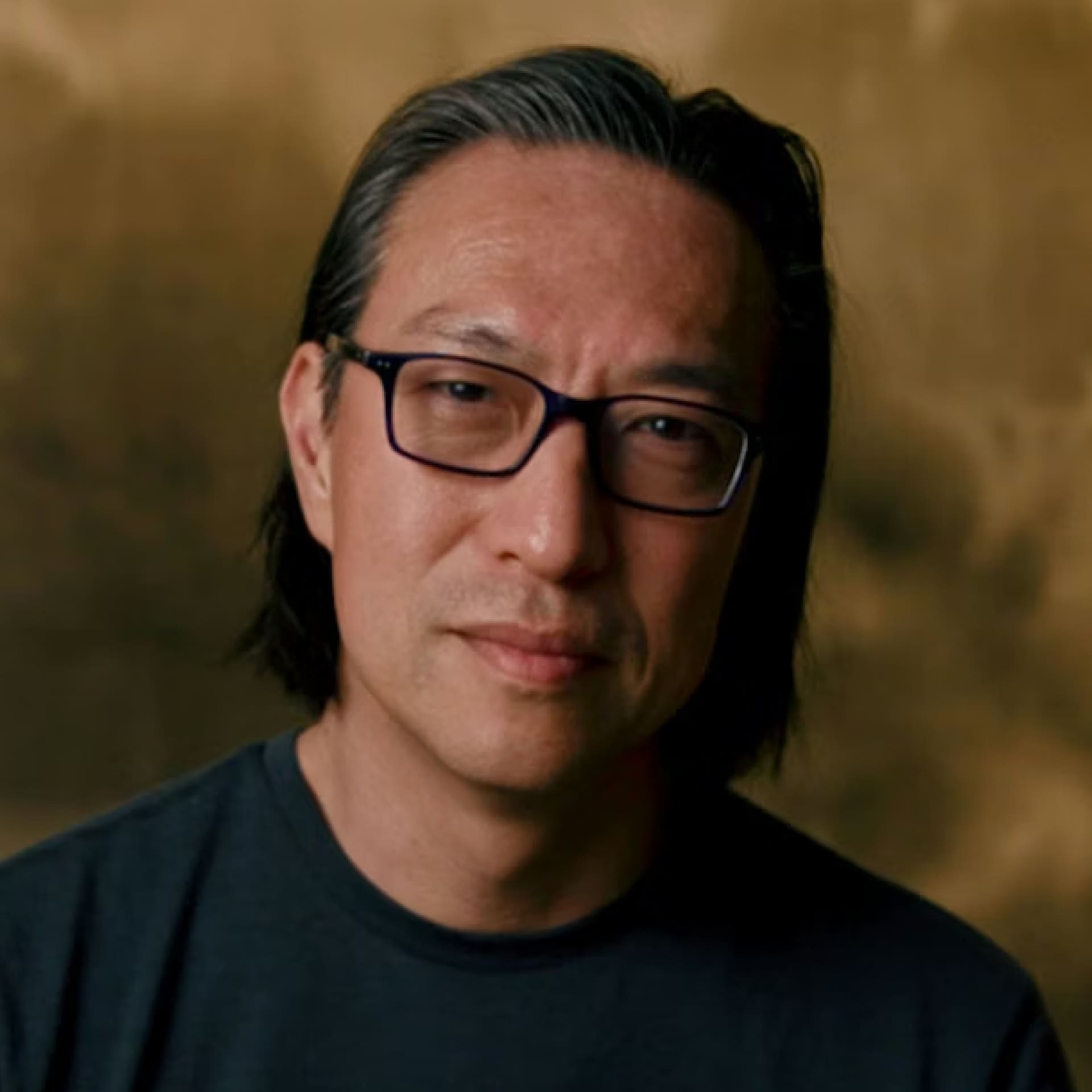 A man with straight, shoulder-length dark hair and glasses, wearing a dark shirt, looks at the camera with a neutral expression against a brown, blurred background.
