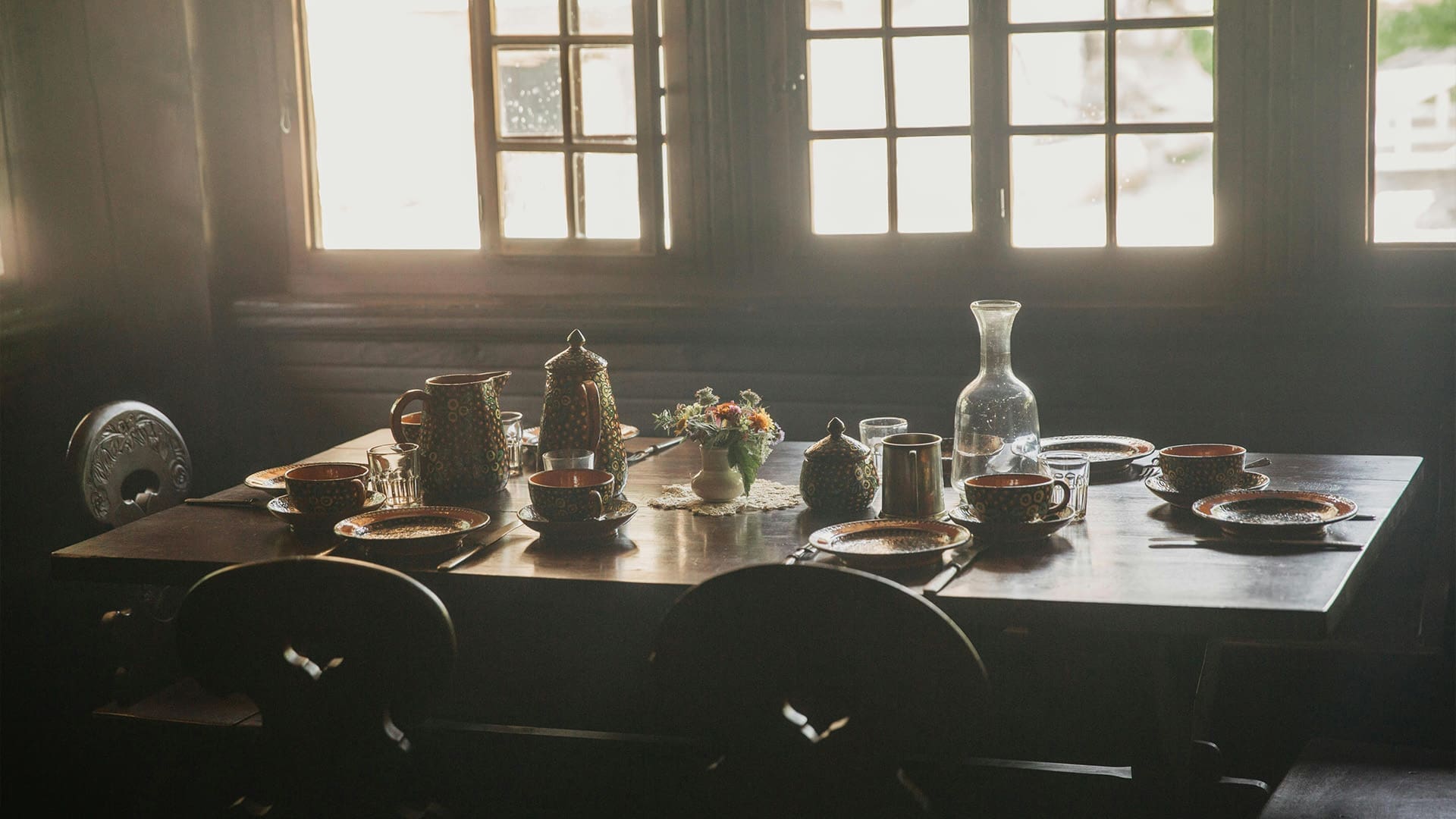 A dark wooden dining table set for four with plates, cups, and a teapot, arranged around a floral centerpiece. Soft light filters in through large windows in the background, creating a cozy atmosphere.
