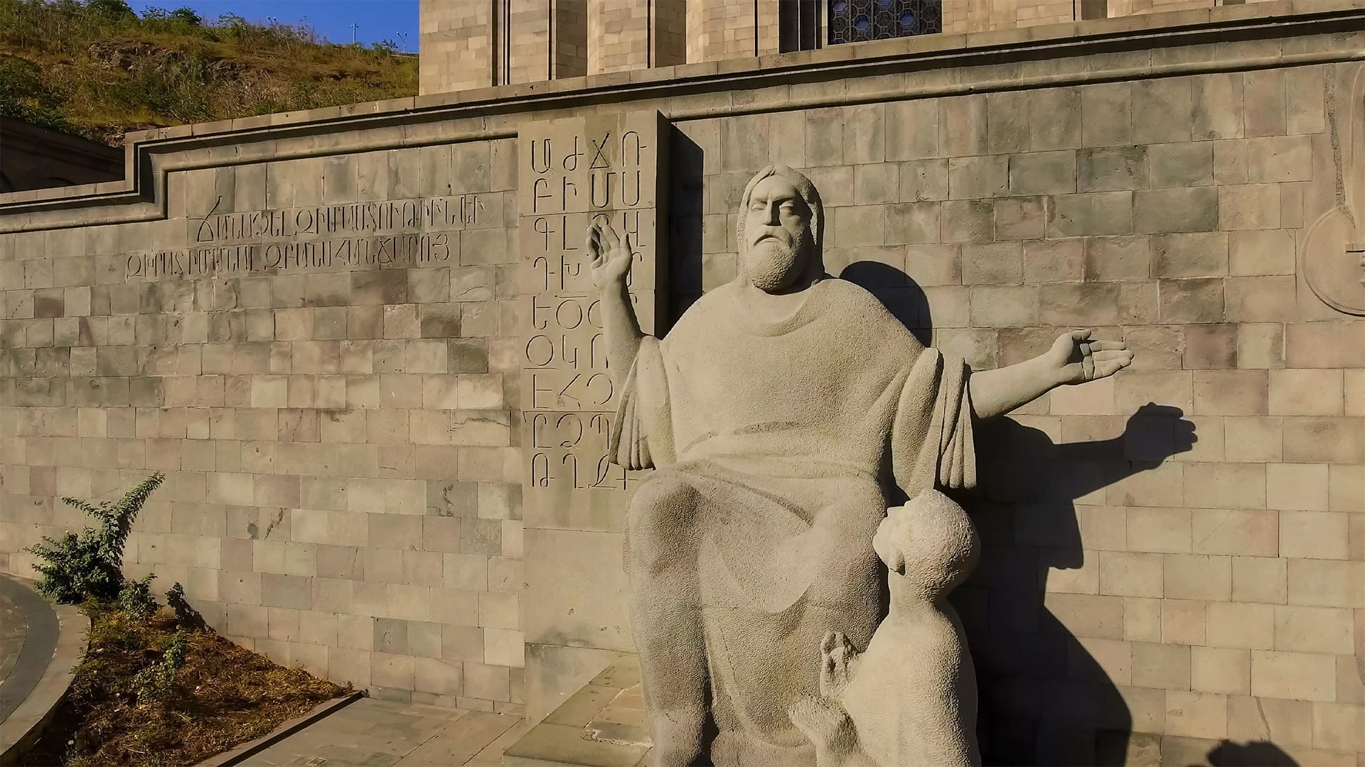 A stone statue of a seated man with a child at his feet is in front of a wall engraved with Armenian script and the Armenian alphabet, outside a gray stone building with stairs and grass nearby.
