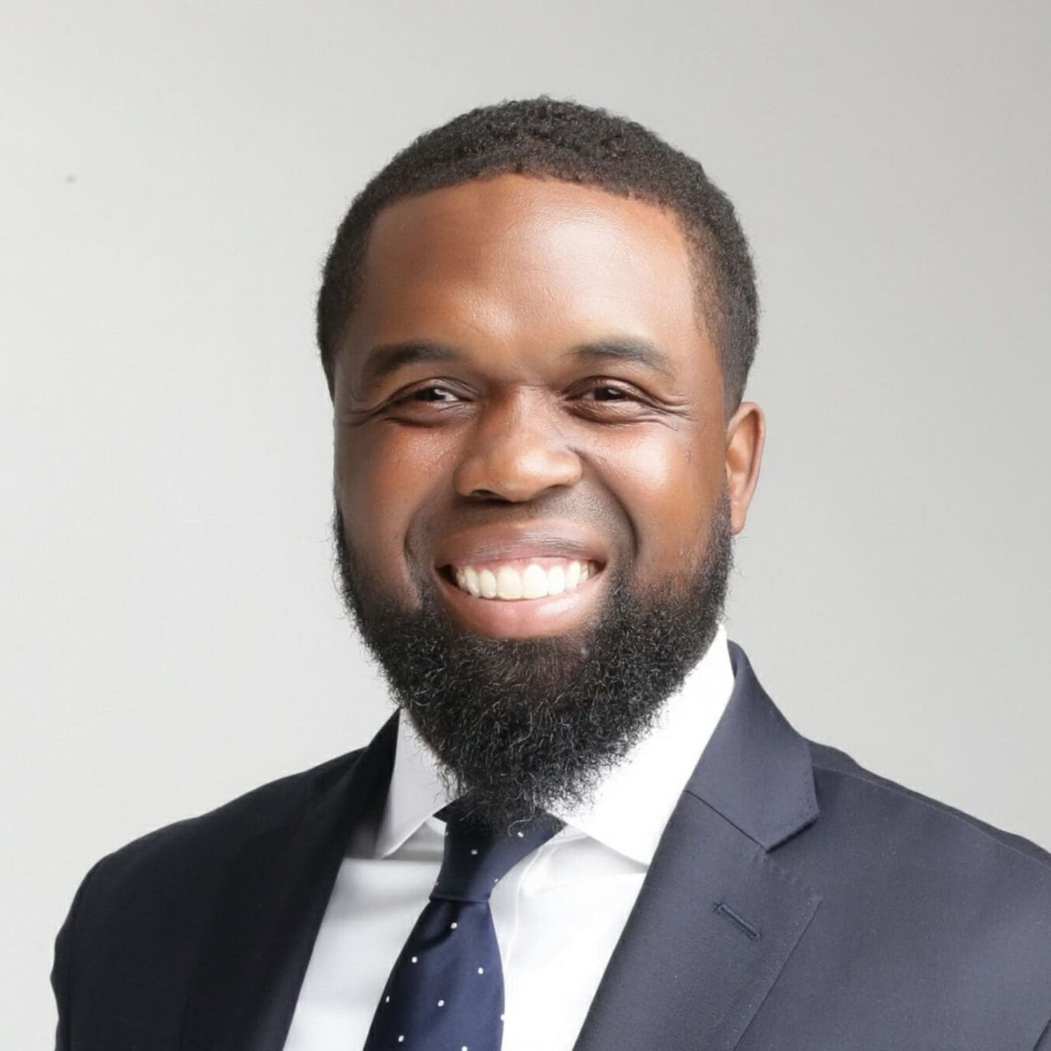 A man with a neatly trimmed beard and short hair smiles warmly while wearing a dark suit, white shirt, and dark polka-dot tie against a plain light background.