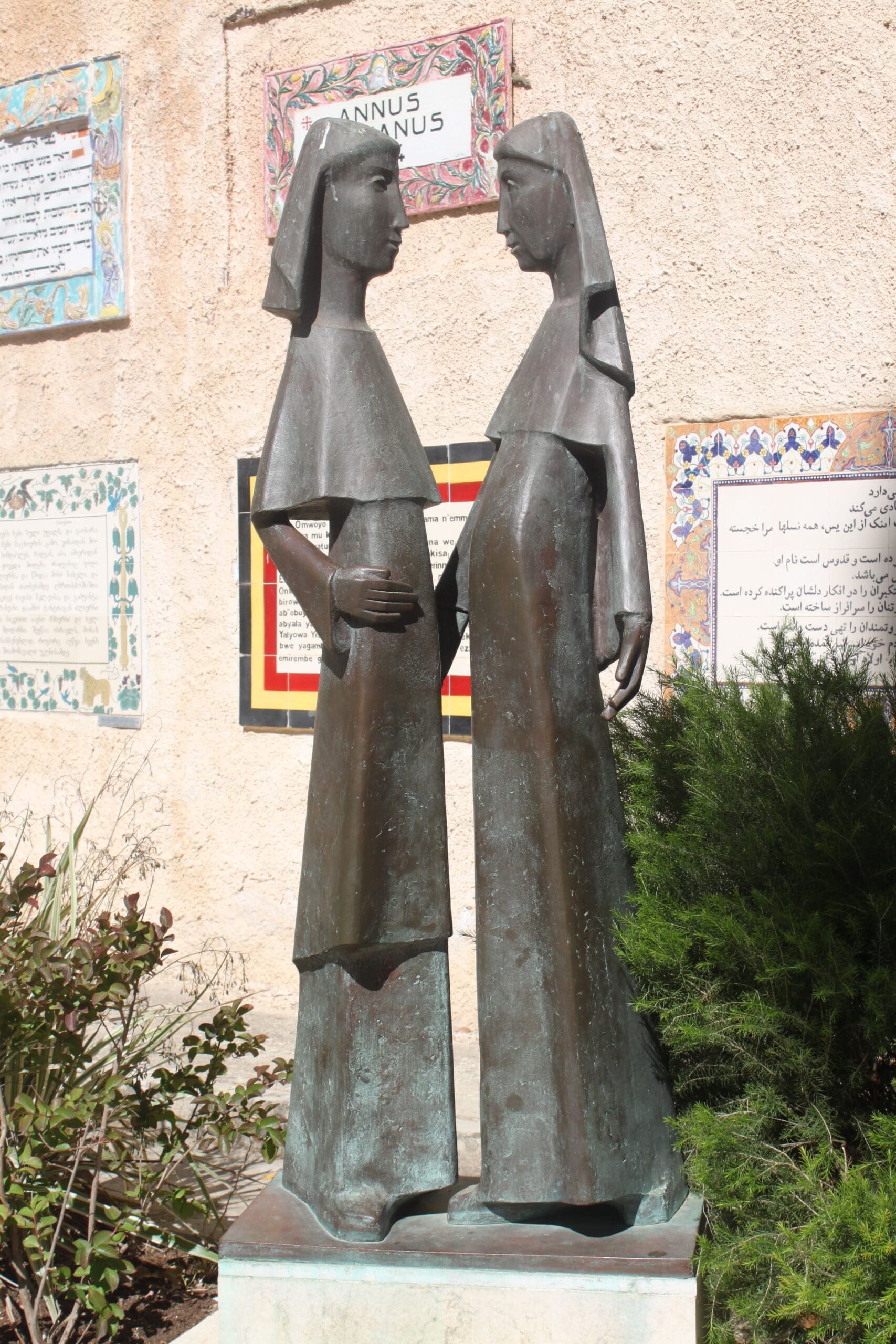 Bronze statue of two robed women standing face-to-face, one with a hand on her belly, against a wall with colorful tiled plaques featuring texts in different languages. Shrubs surround the statues base.