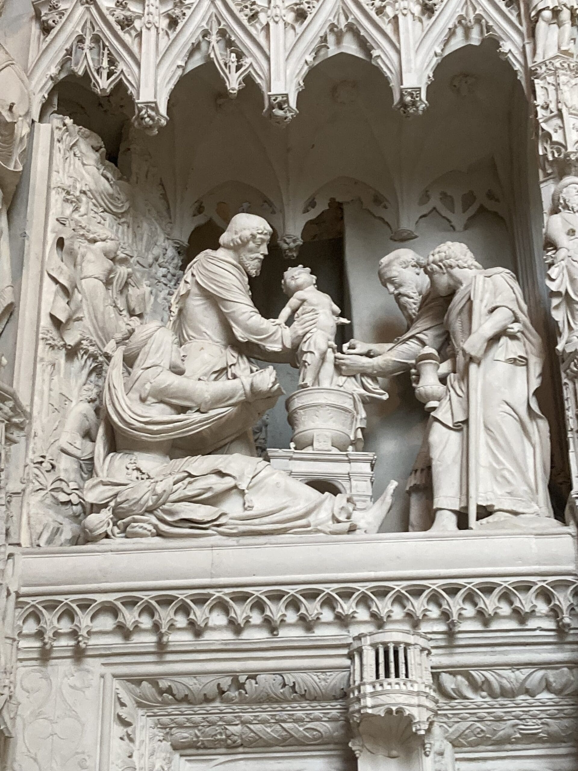 A detailed marble relief shows four adults and a baby, with one adult holding the child above a basin, likely depicting a baptism, set in a gothic architectural frame with ornate carvings.