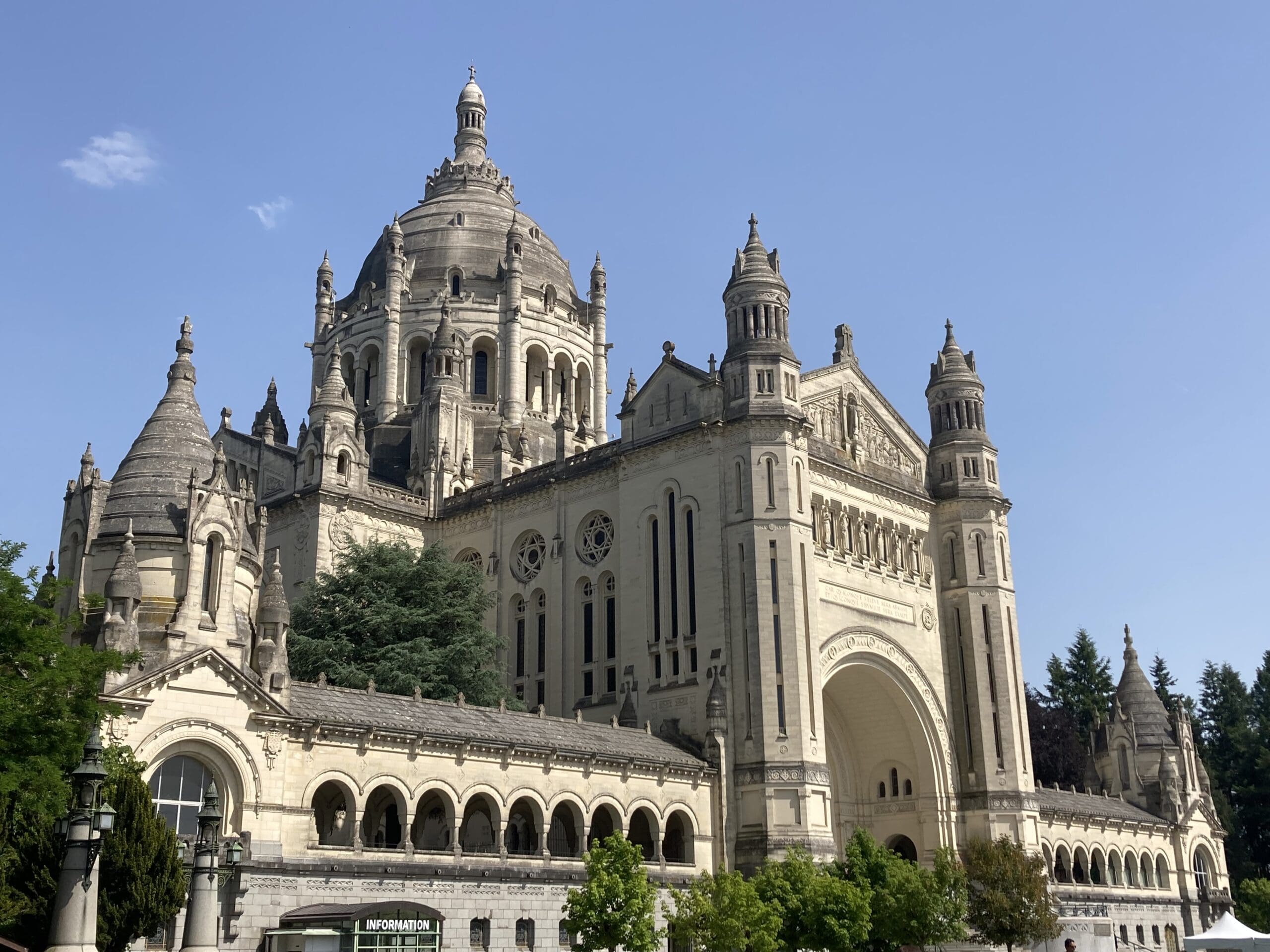 Large stone basilica with ornate towers, domes, and arches under a clear blue sky, surrounded by green trees and a few people near the entrance.