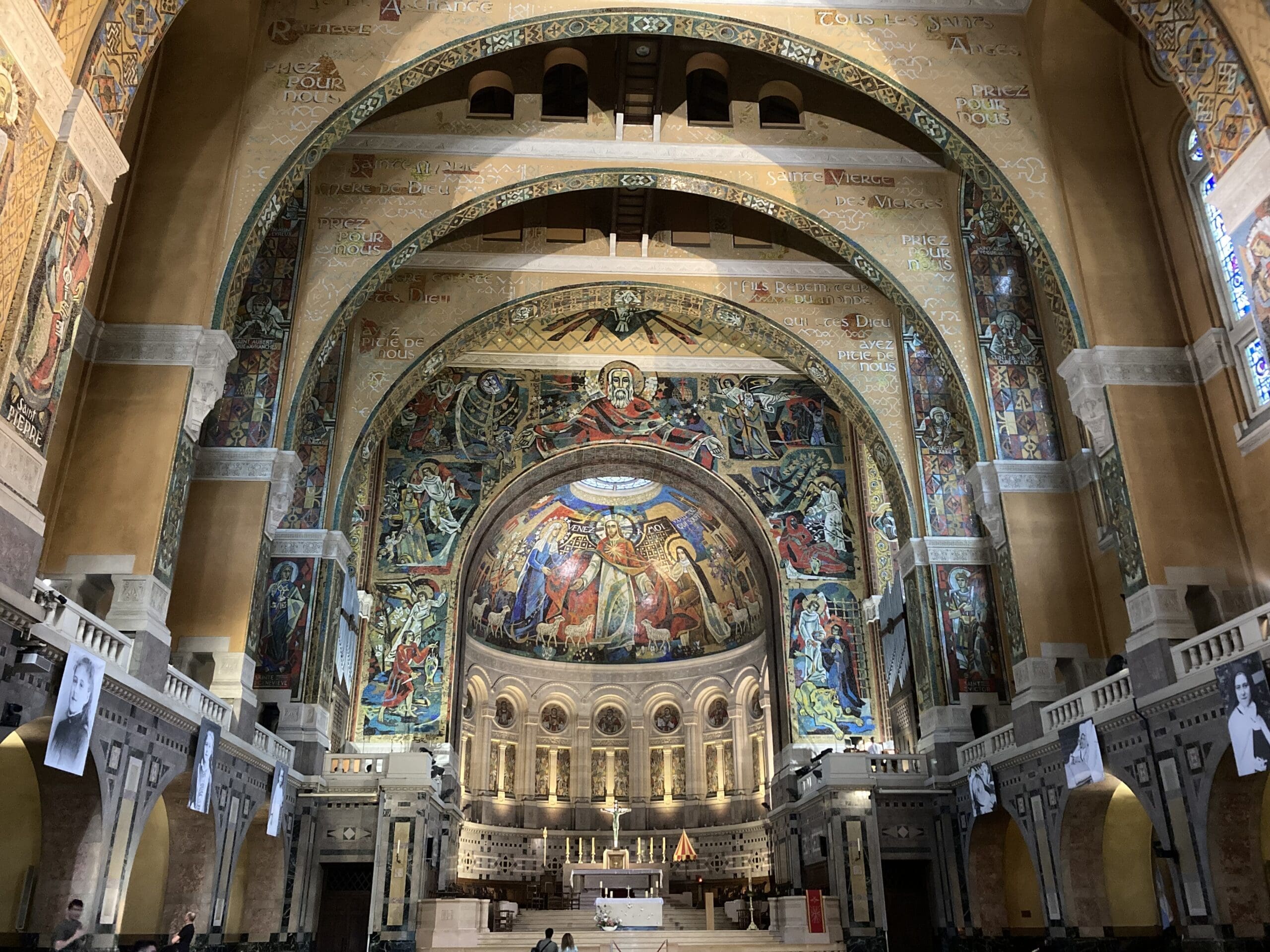 Interior of a grand church with colorful mosaics and paintings on arched ceilings and walls, ornate columns, and banners with black-and-white portraits hanging along the sides. The altar is visible at the center.