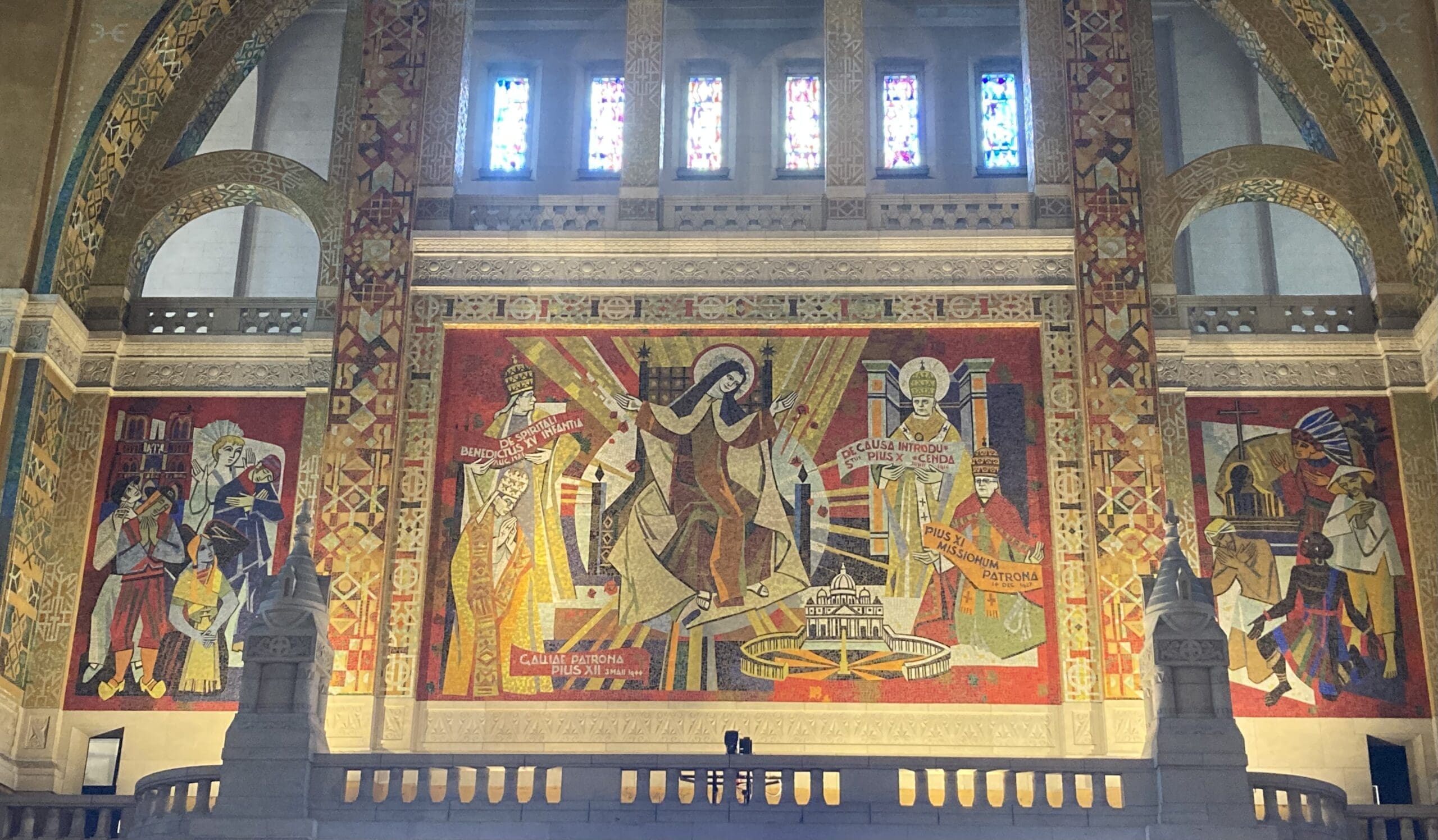 Colorful mosaic mural in a cathedral, featuring a central figure with arms raised and two other figures on either side, with stained glass windows above, and ornate architectural details.