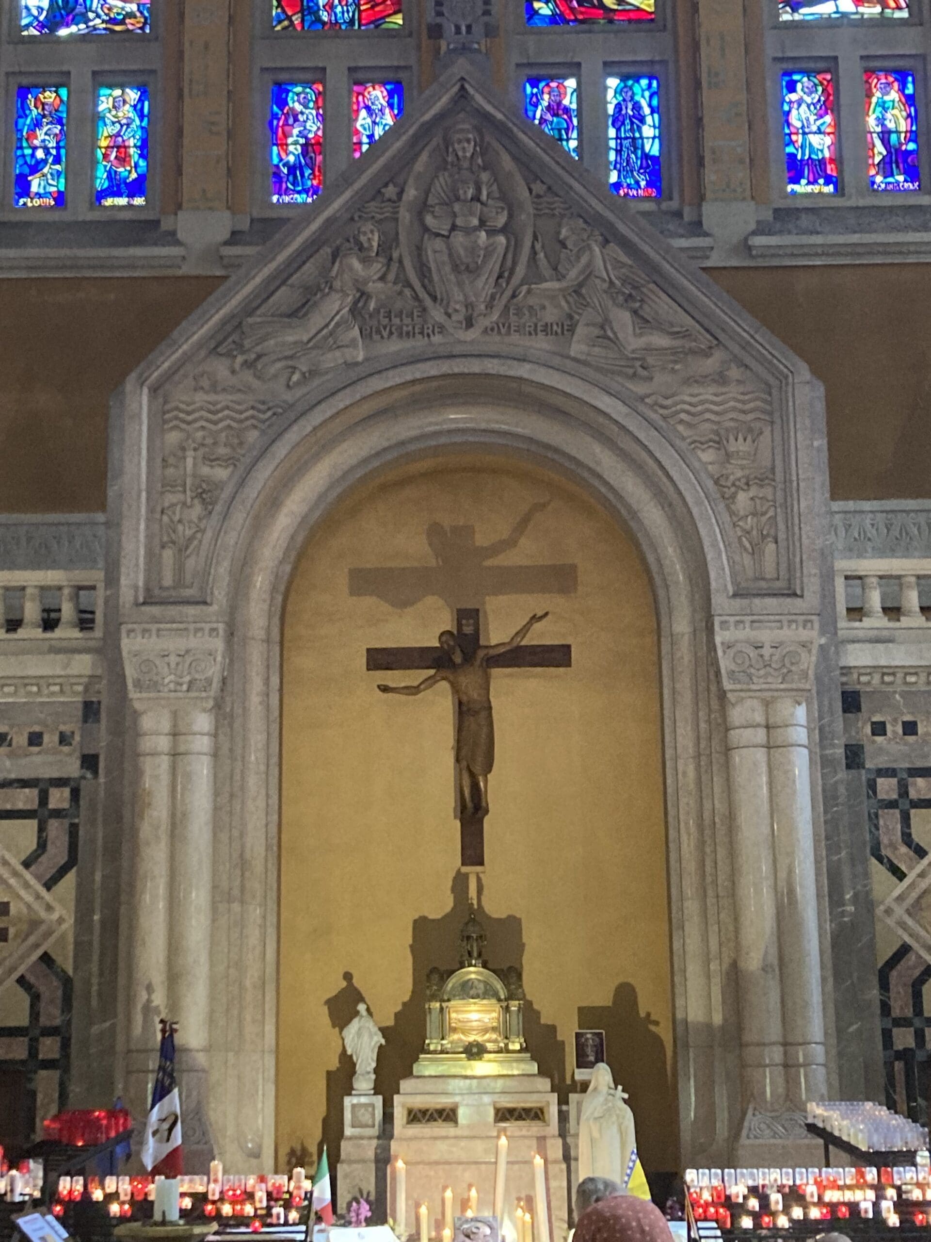 Altar in a church with a crucifix, candles, and two statues. Stained glass windows are above, and the crucifixs shadow is cast on the wall behind. The scene is ornate and solemn.