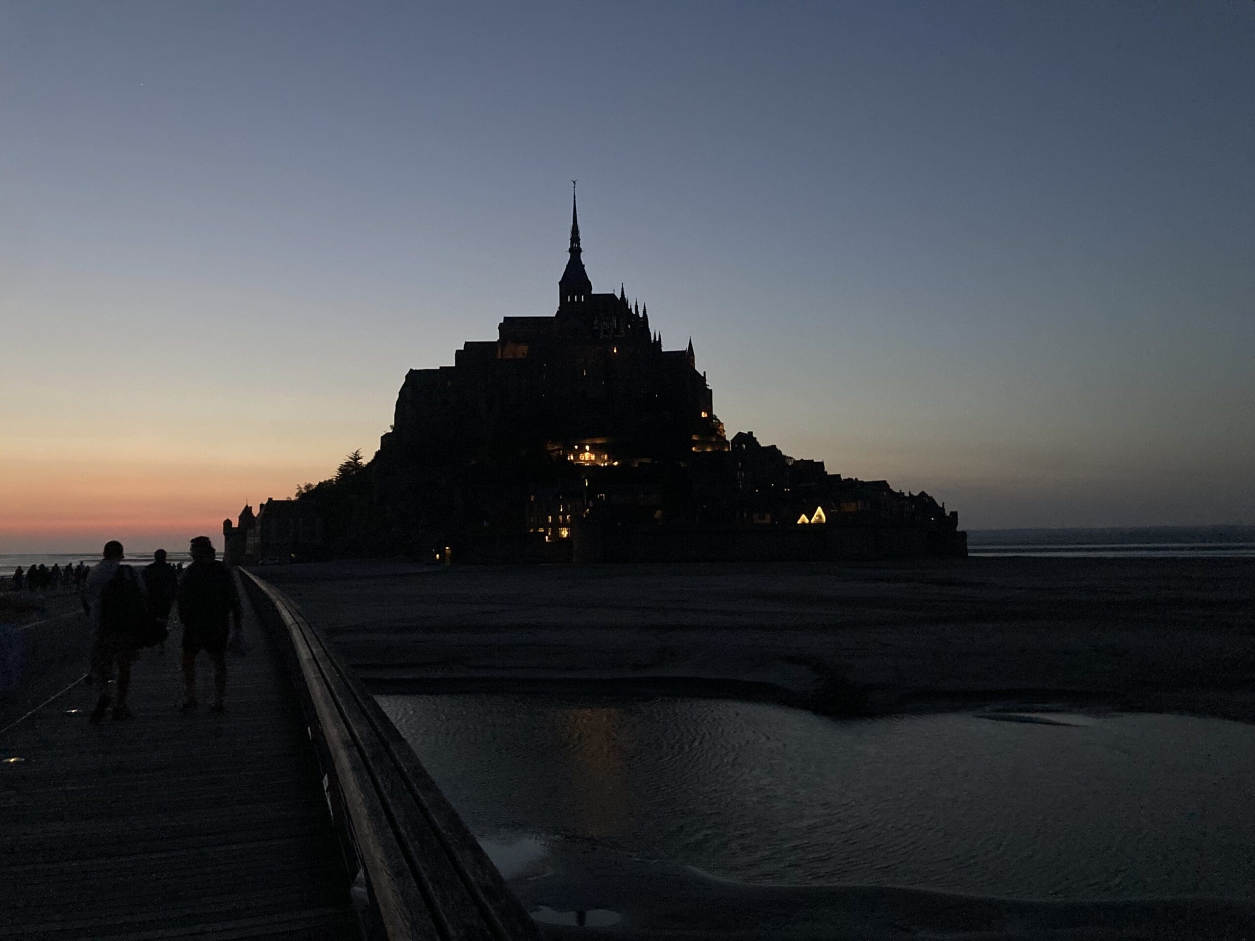 Silhouette of Mont Saint-Michel at dusk, with a few lights glowing in the buildings. The sky is darkening, showing hues of orange and blue, and a walkway leads toward the island over calm water.