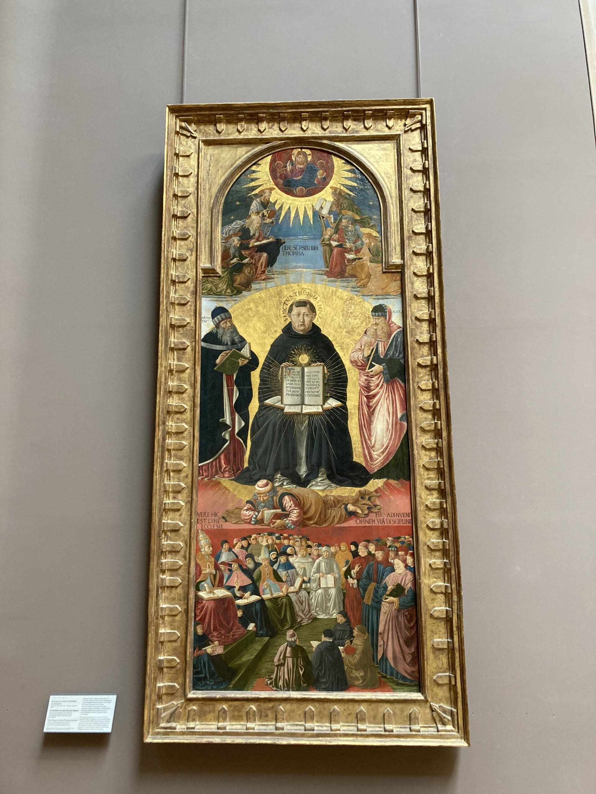 A framed religious painting featuring a central seated figure holding an open book, surrounded by smaller figures above and below, with gold detailing and a decorative border.