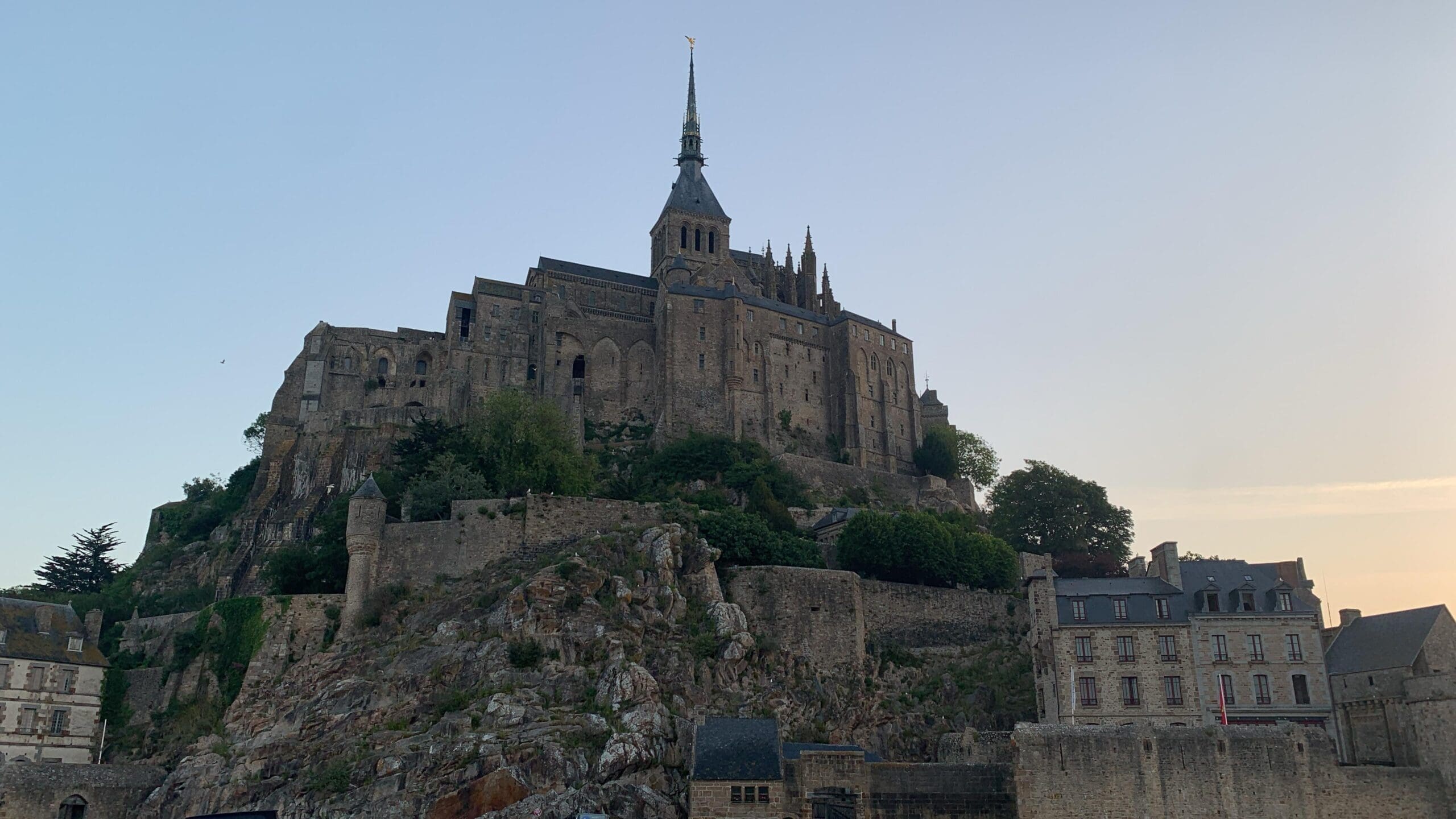 Historic stone abbey atop a rocky hill, surrounded by walls and smaller stone buildings, with a tall spire reaching into the sky at sunset. The scene is peaceful and bathed in soft light.