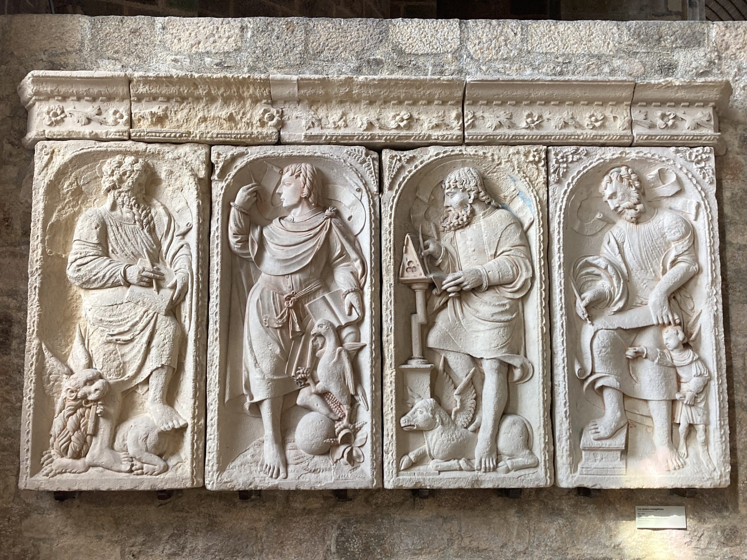 Stone relief with four figures in robes, each with different poses and symbolic objects; animals at their feet and decorative arches above each figure. The relief has intricate details and a weathered appearance.