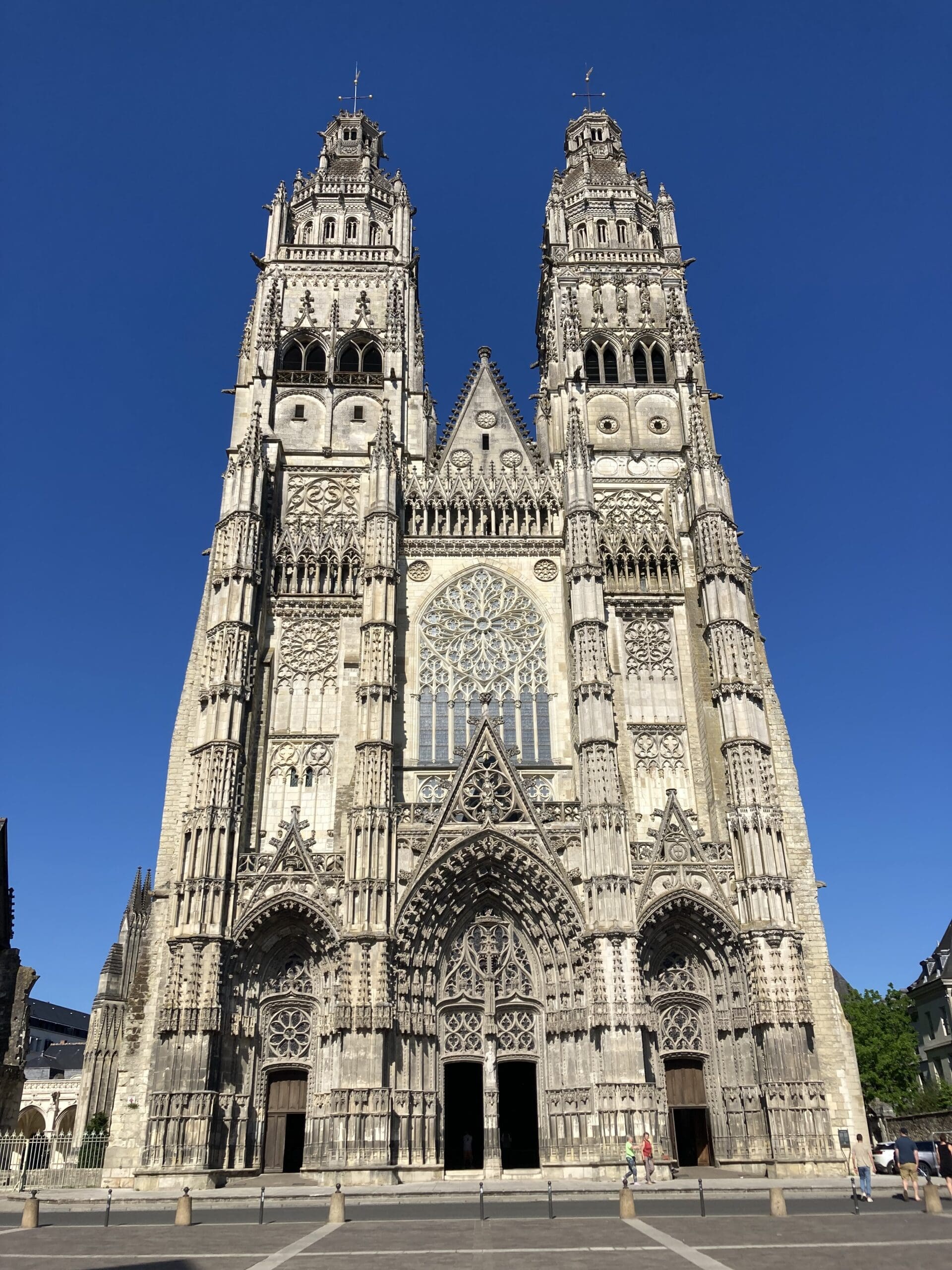 Gothic-style cathedral with two tall, ornate towers, intricate stone carvings, and large stained-glass windows, set against a clear blue sky. A few people are visible near the entrance.
