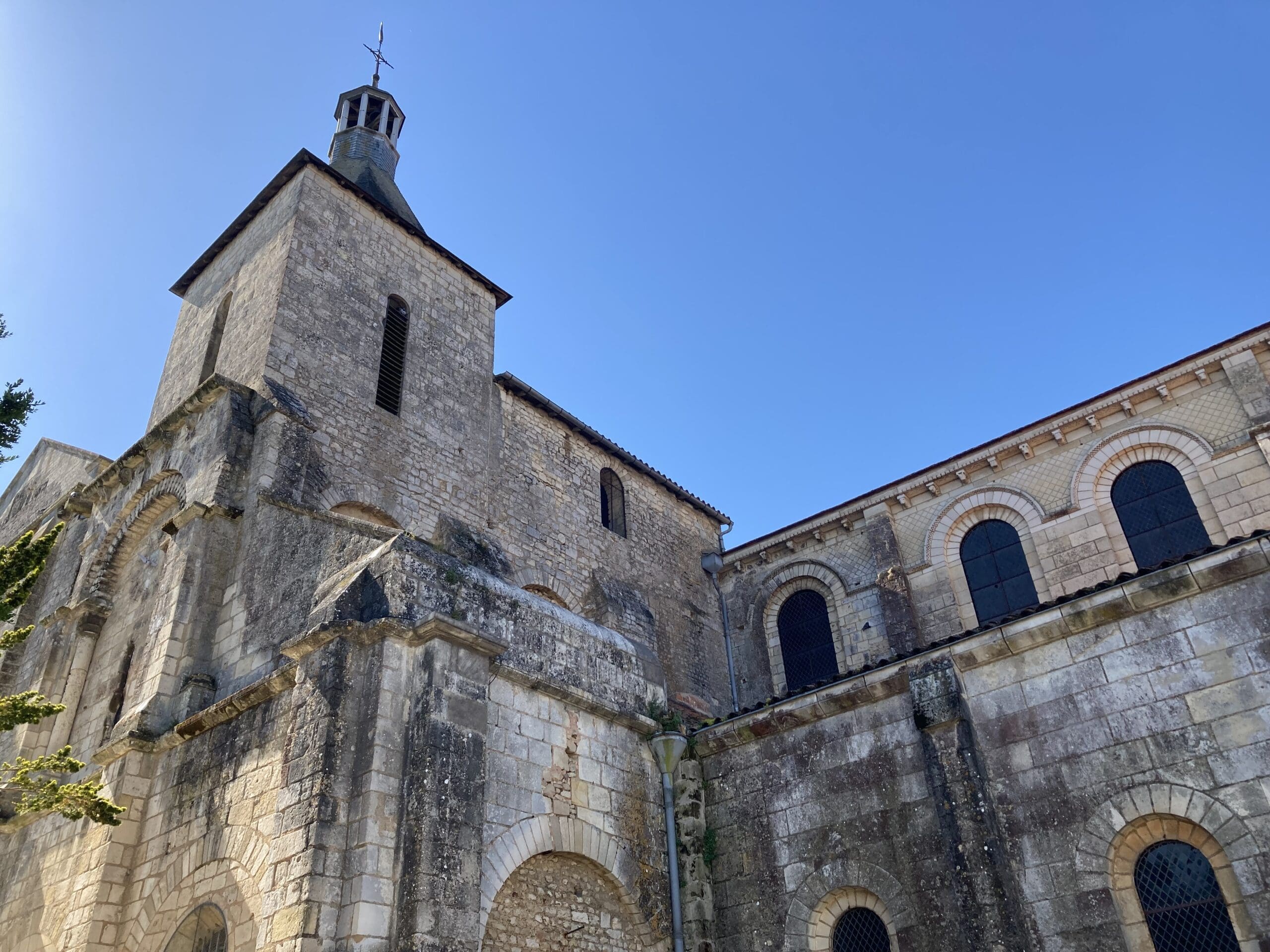 Stone church with arched windows and a bell tower set against a clear blue sky, seen from a low angle. The building has weathered stone walls and some greenery is visible on the left.
