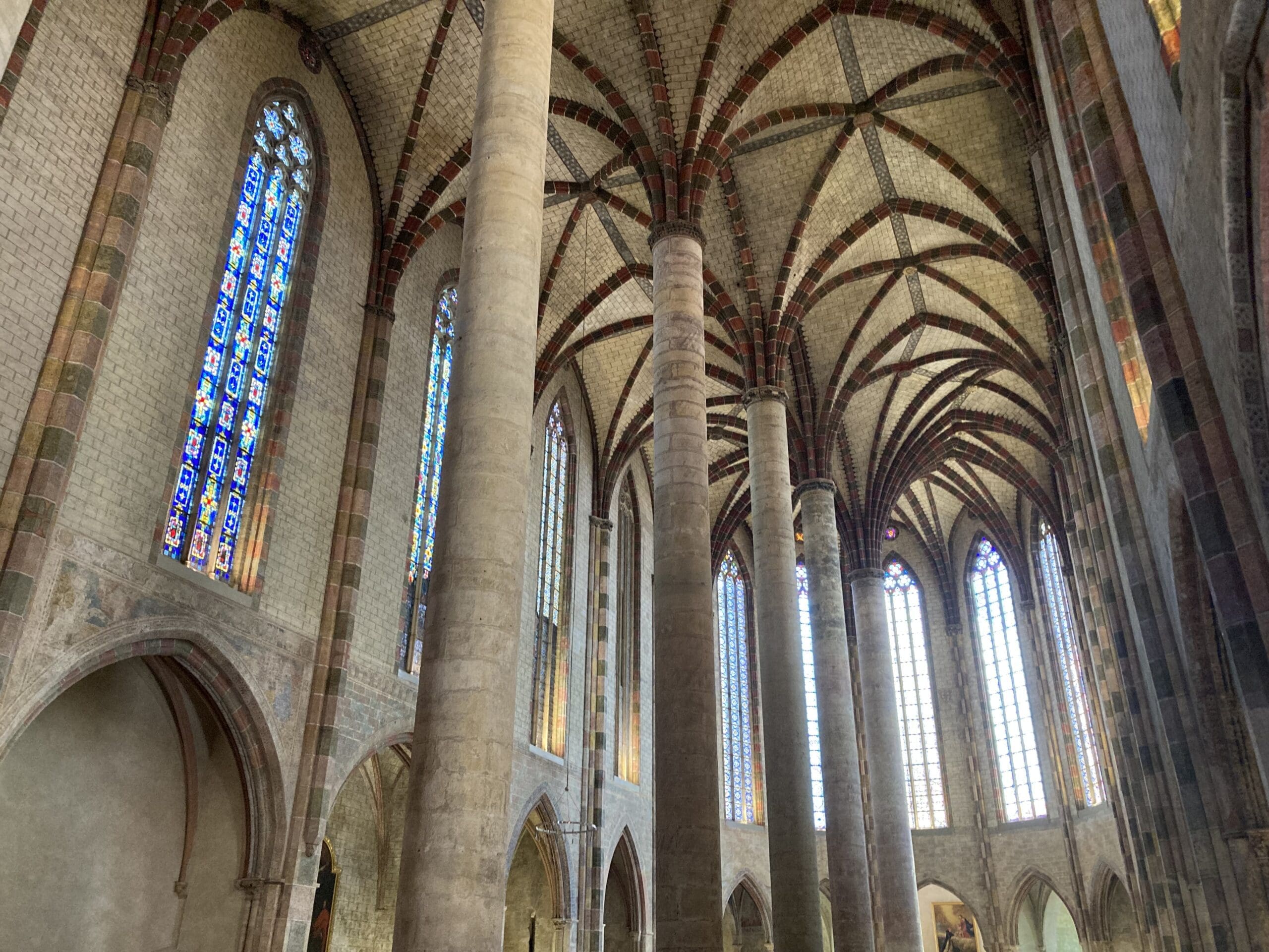Interior of a Gothic cathedral with tall stone columns, stained glass windows, and an intricate ribbed vaulted ceiling, illuminated by natural light.