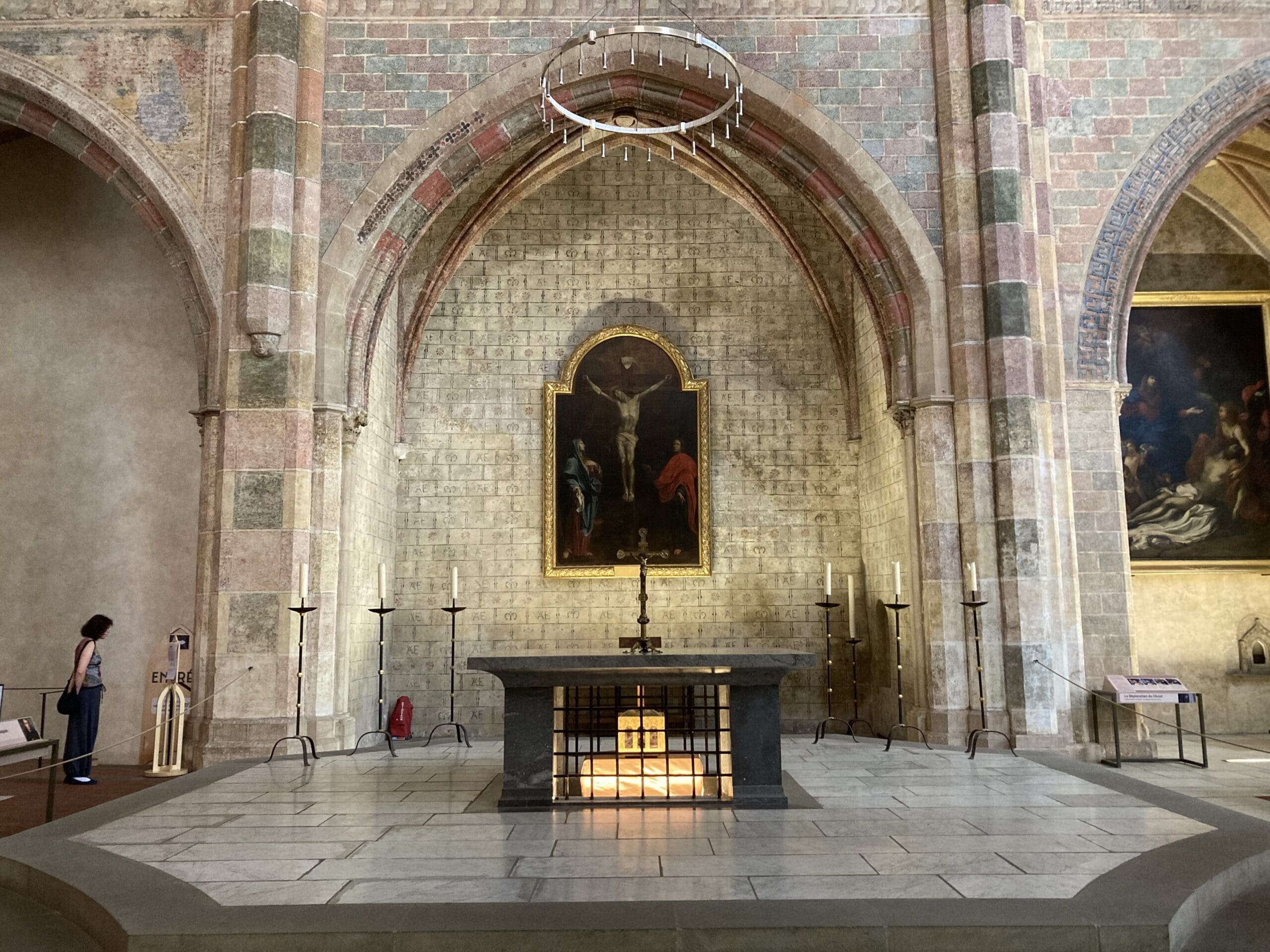 A stone altar sits beneath a circular chandelier and a religious painting in a high-arched church interior; a person stands to the left, observing the scene.