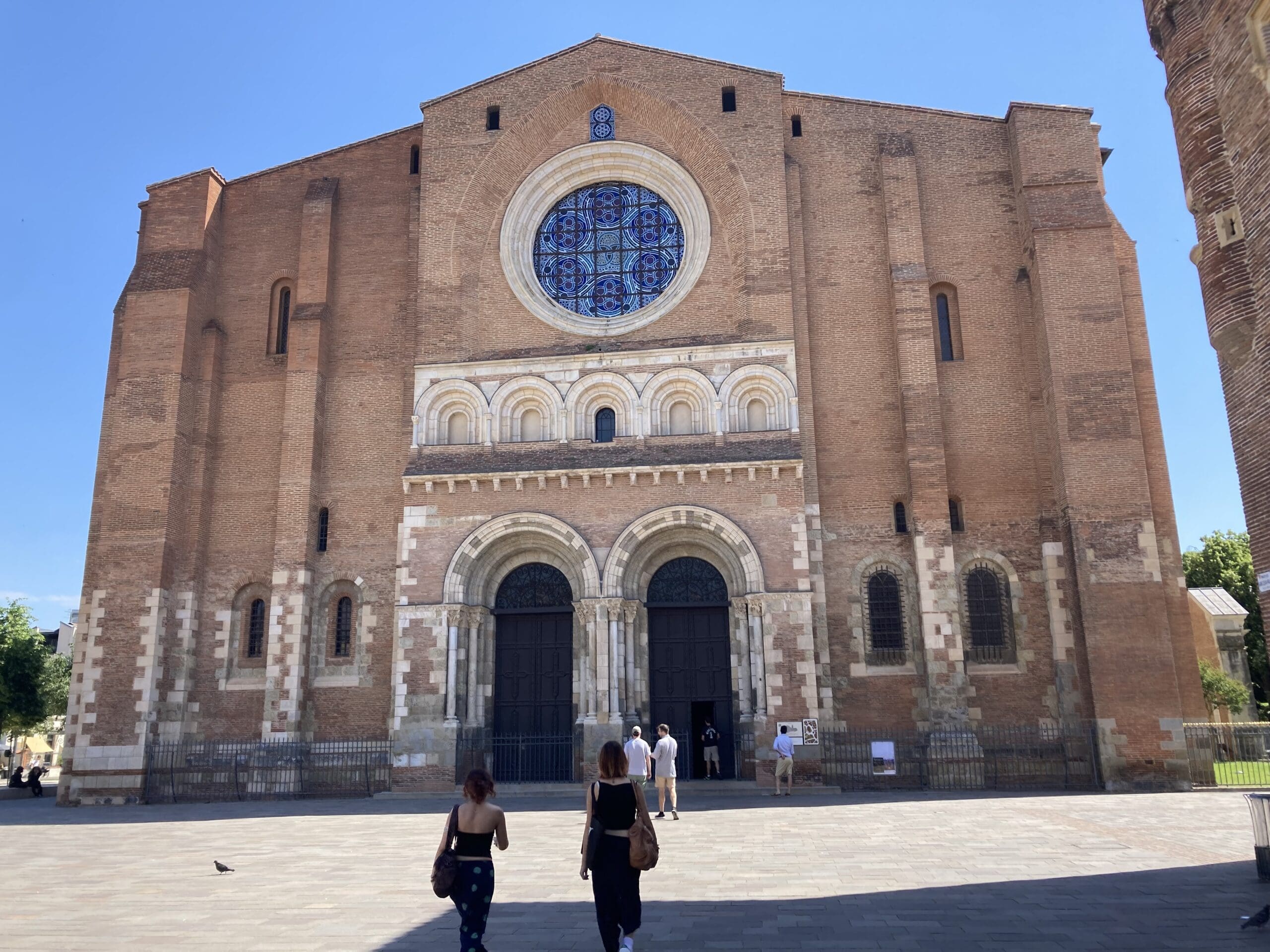 People walk toward a large brick church with arched doorways, a circular stained glass window, and decorative stonework on a sunny day.