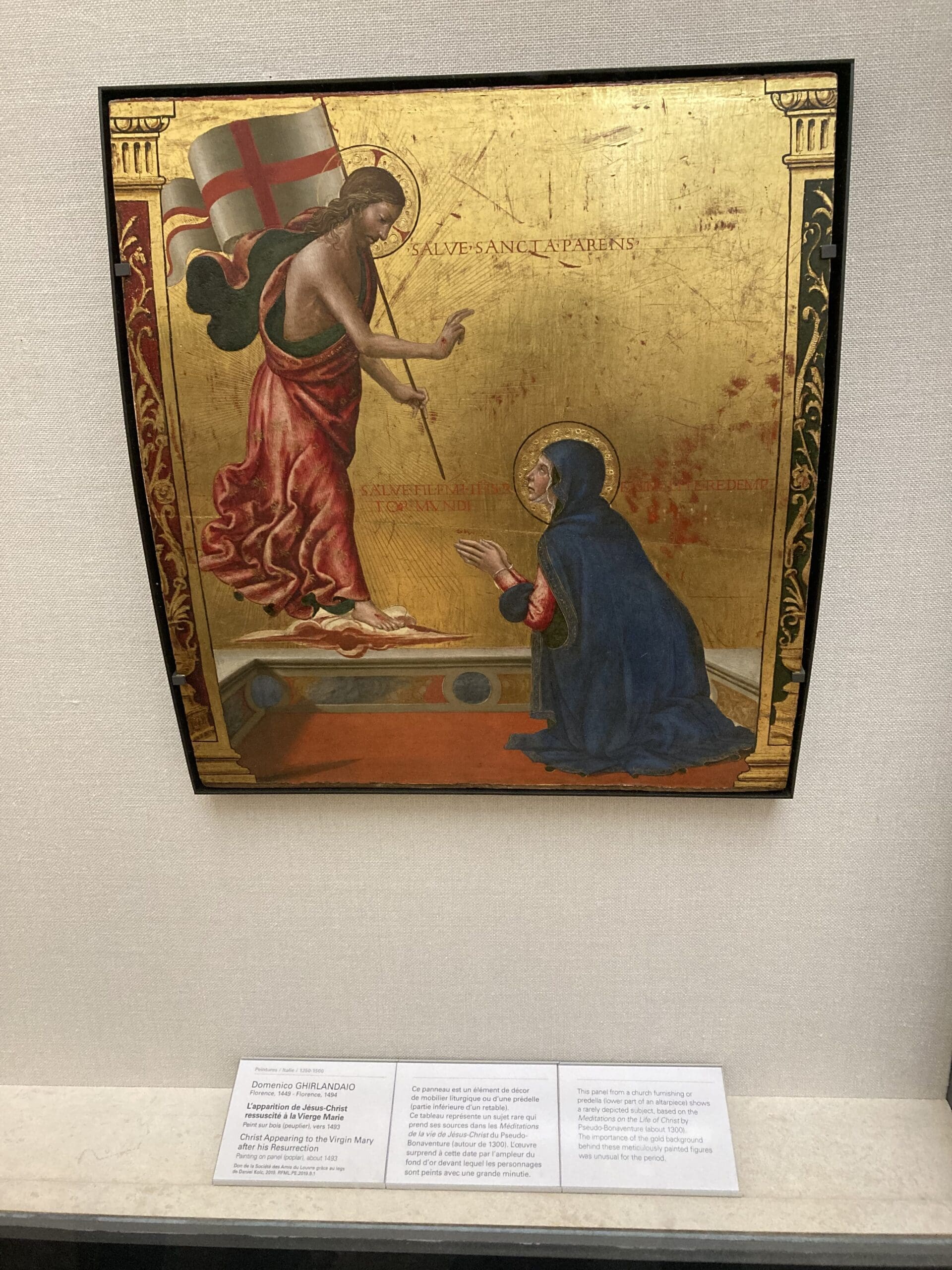 A gold-leaf religious painting shows a haloed, robed figure with a staff and flag appearing to a kneeling woman in blue robes, who has a gold halo. Descriptive plaques are displayed below the artwork.