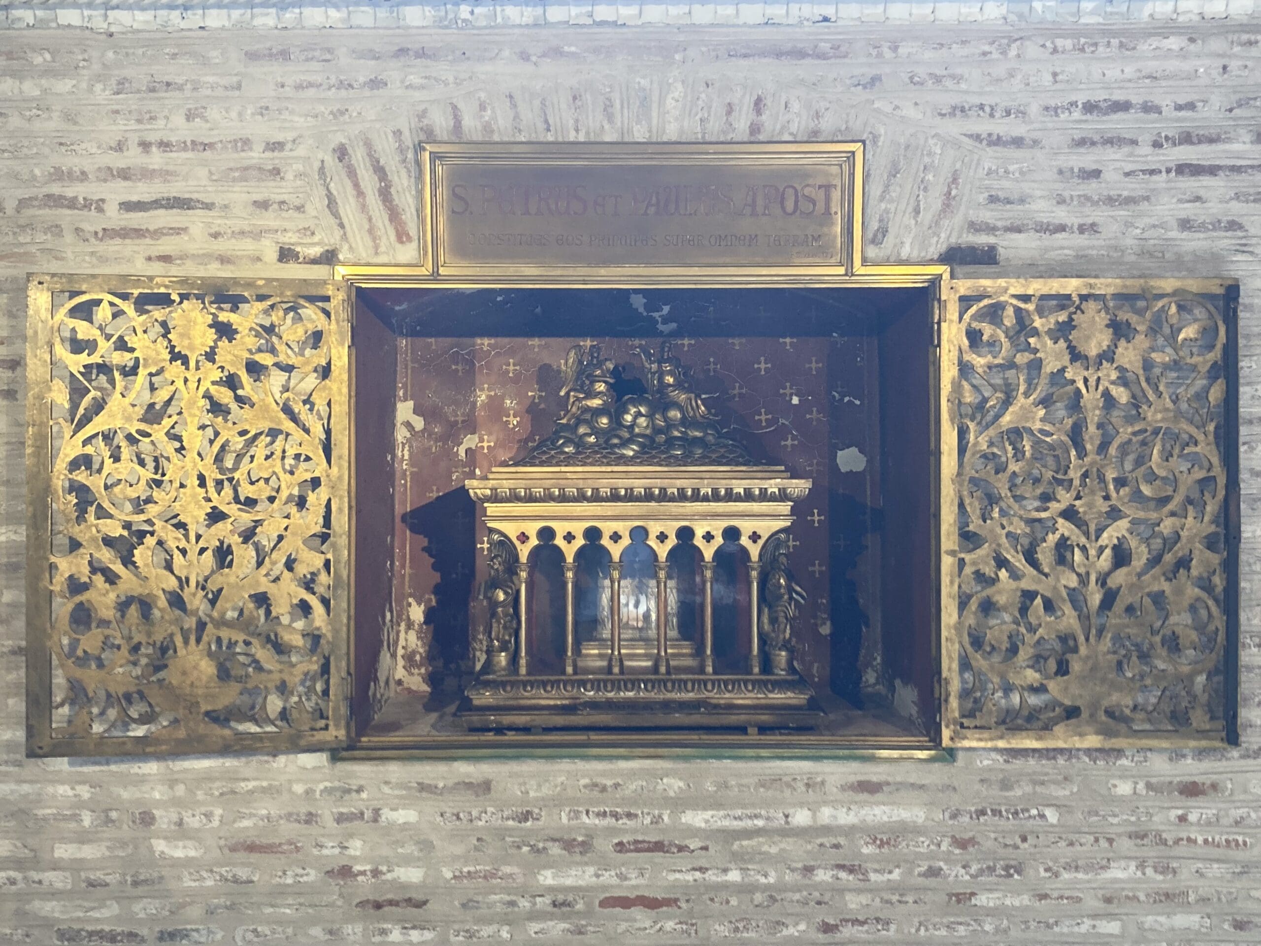 A golden ornate reliquary with intricate doors is set in a brick wall alcove, displayed behind glass, with a Latin inscription above it.