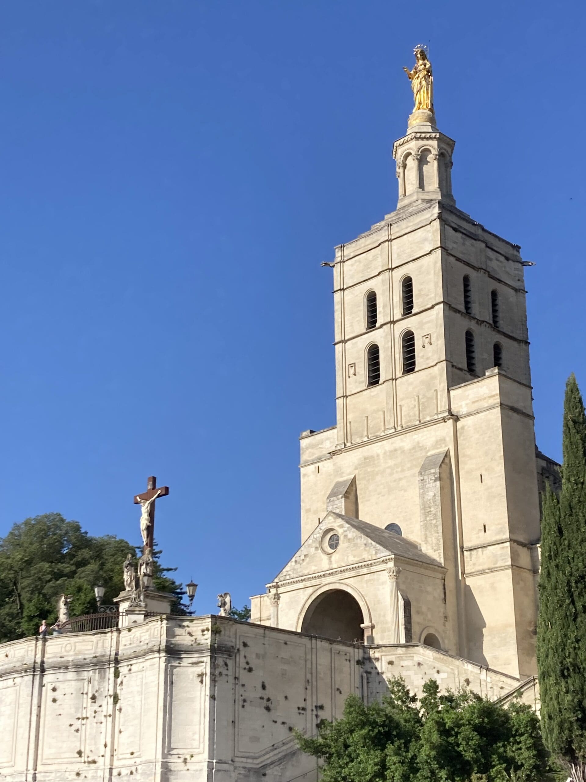 A tall stone church with a prominent bell tower topped by a gold statue, a cross with a statue in front, and a clear blue sky in the background.