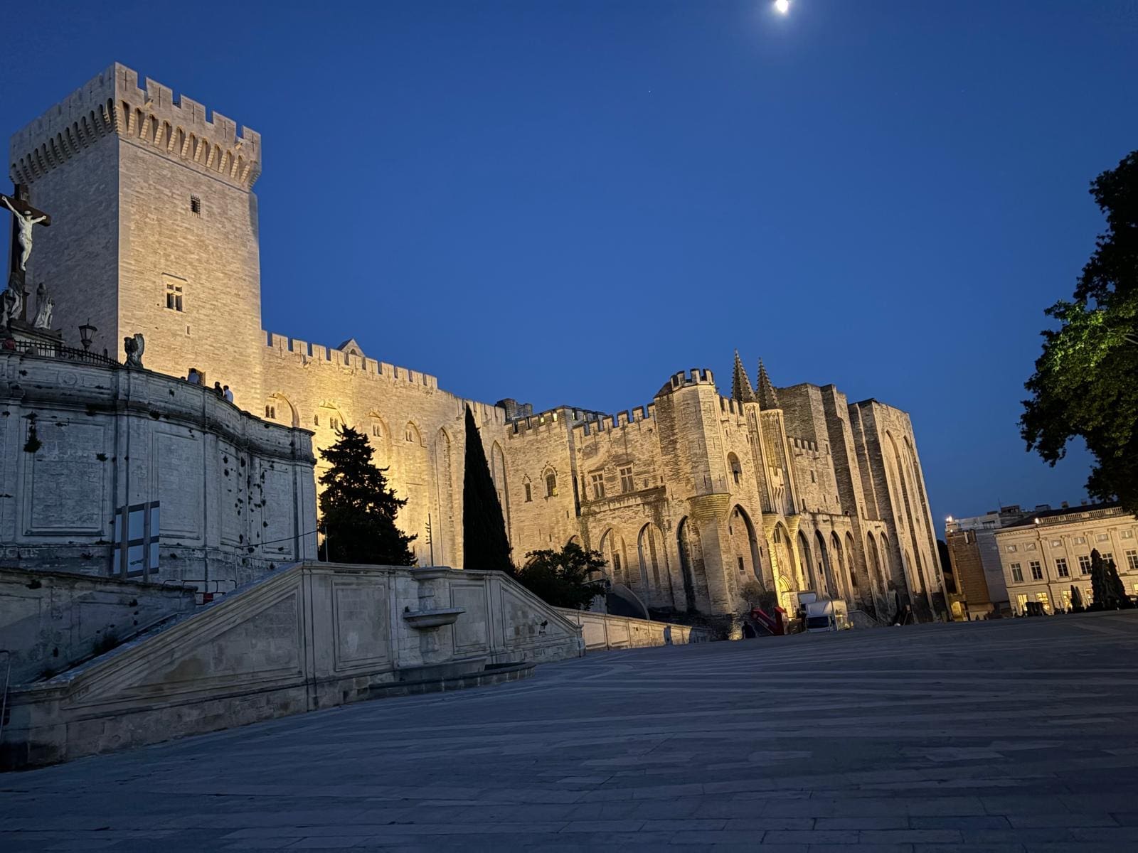 An illuminated medieval stone palace with towers and arches is seen at dusk under a clear sky with a visible moon, and an empty stone plaza in the foreground.