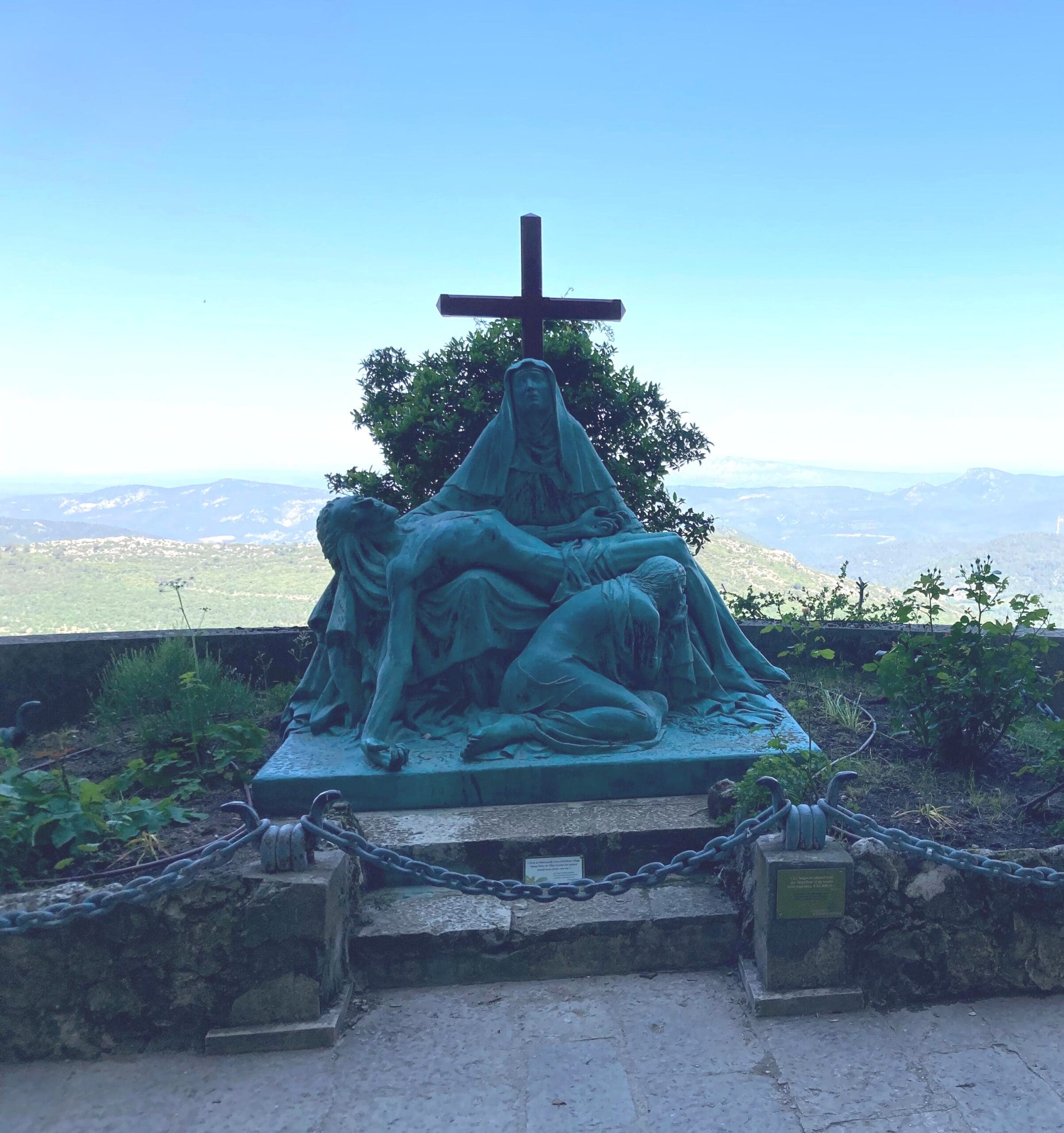 A bronze statue depicts three mournful figures at the base of a cross, overlooking a scenic landscape with mountains and greenery under a clear sky.
