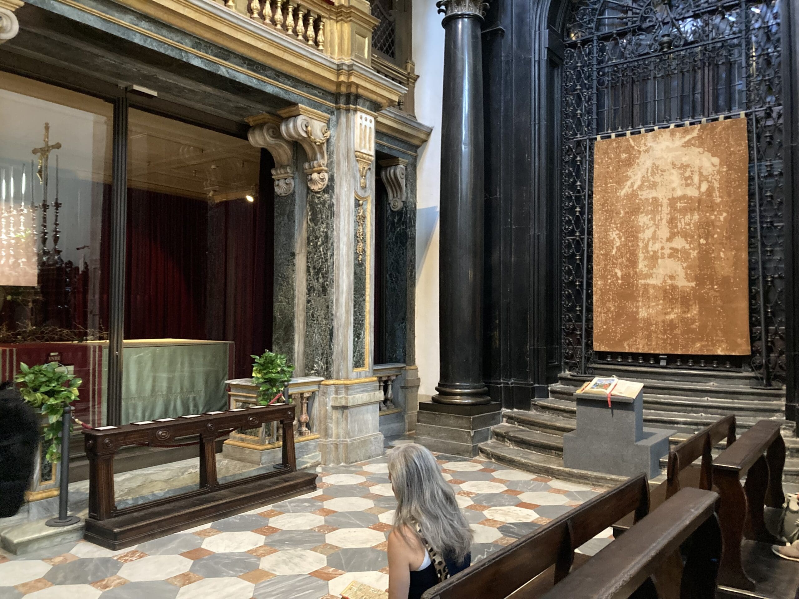 A woman kneels in prayer inside a church, facing a display case and a large cloth with a faint face image, surrounded by ornate columns and marble floors.