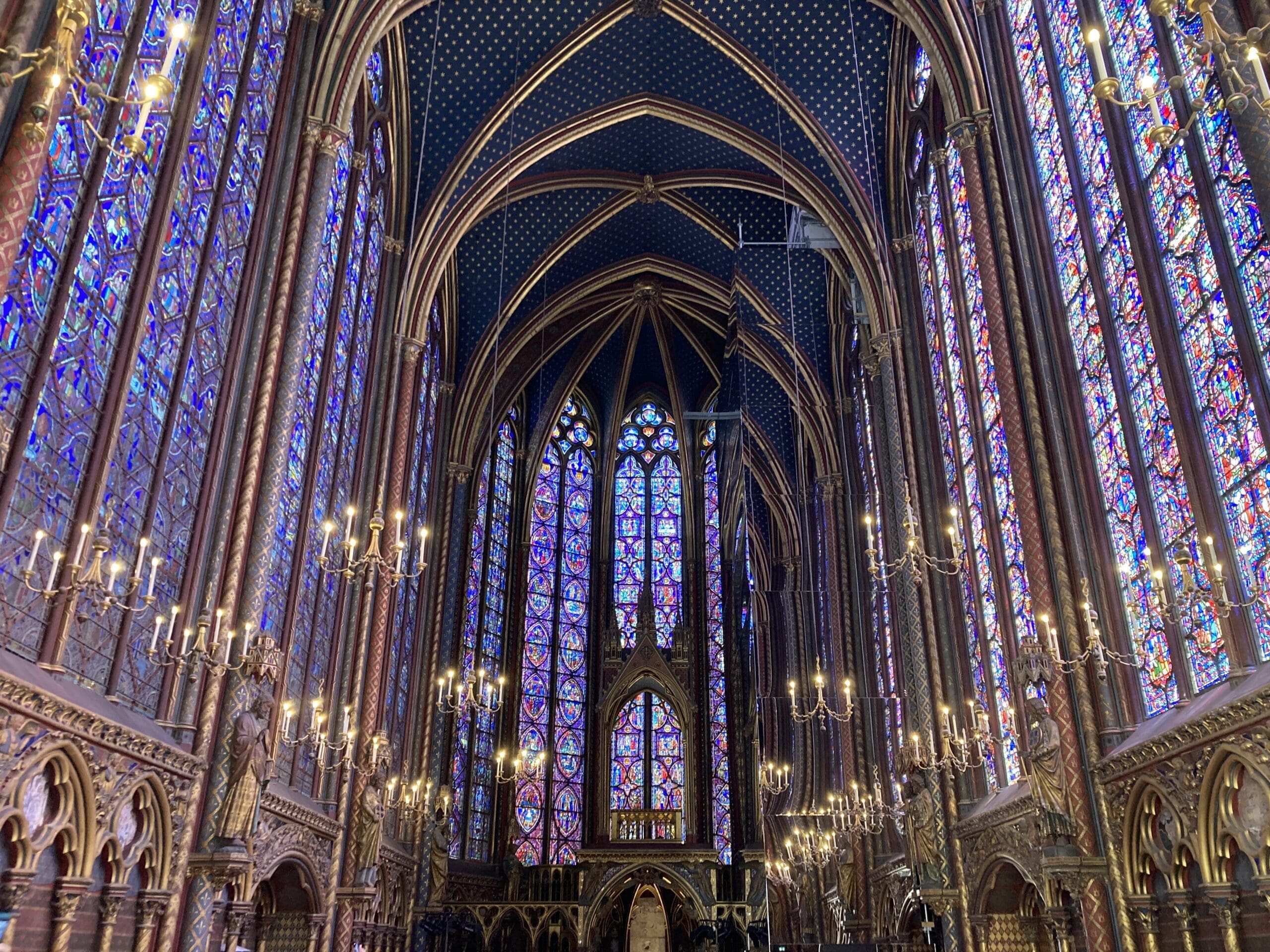 Interior of a Gothic cathedral with high arched ceilings, ornate columns, and vibrant stained glass windows. Chandeliers hang from the ceiling, illuminating detailed carvings and intricate patterns on the walls.