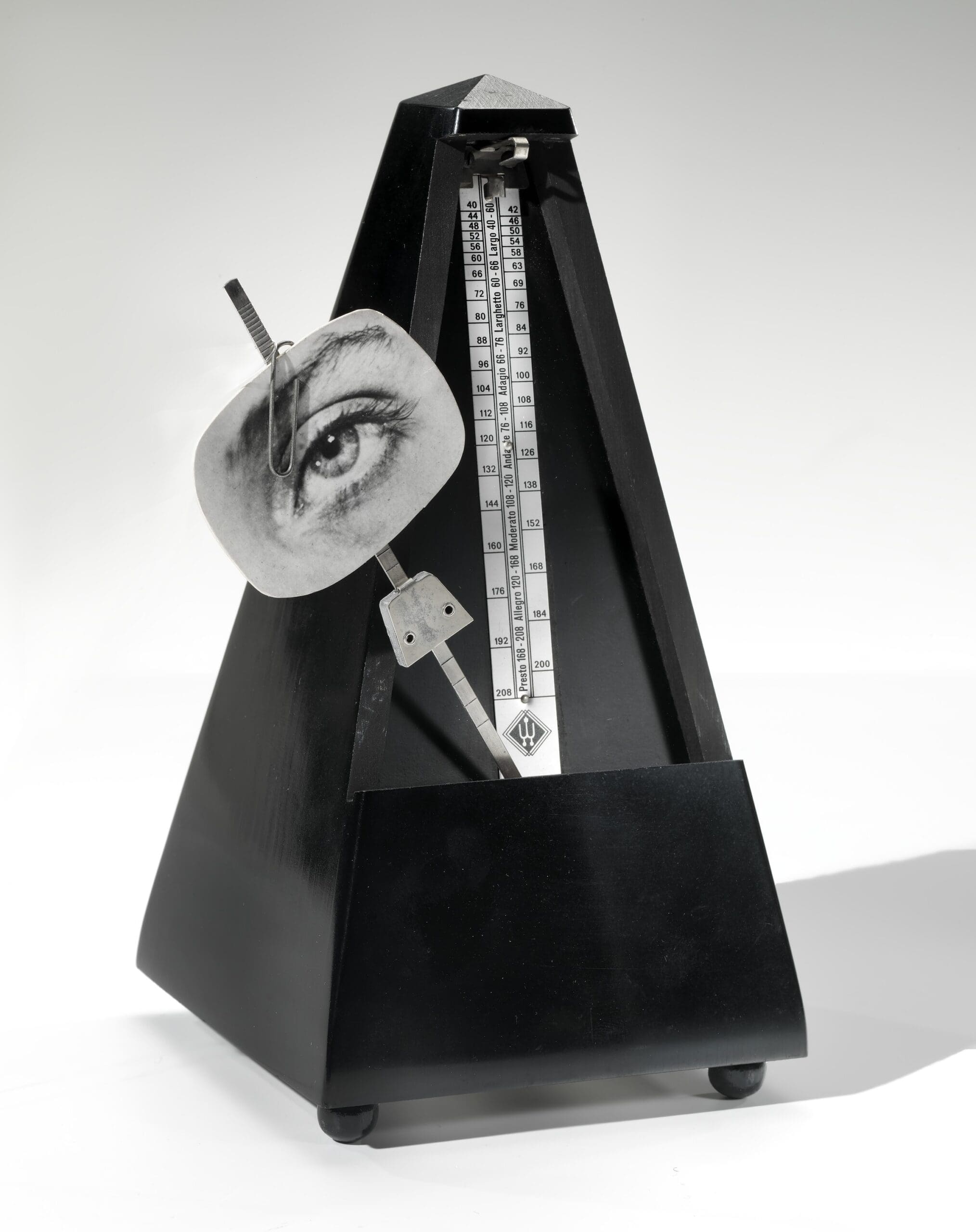A black metronome with a black-and-white cutout of a human eye attached to its pendulum, set against a plain white background.