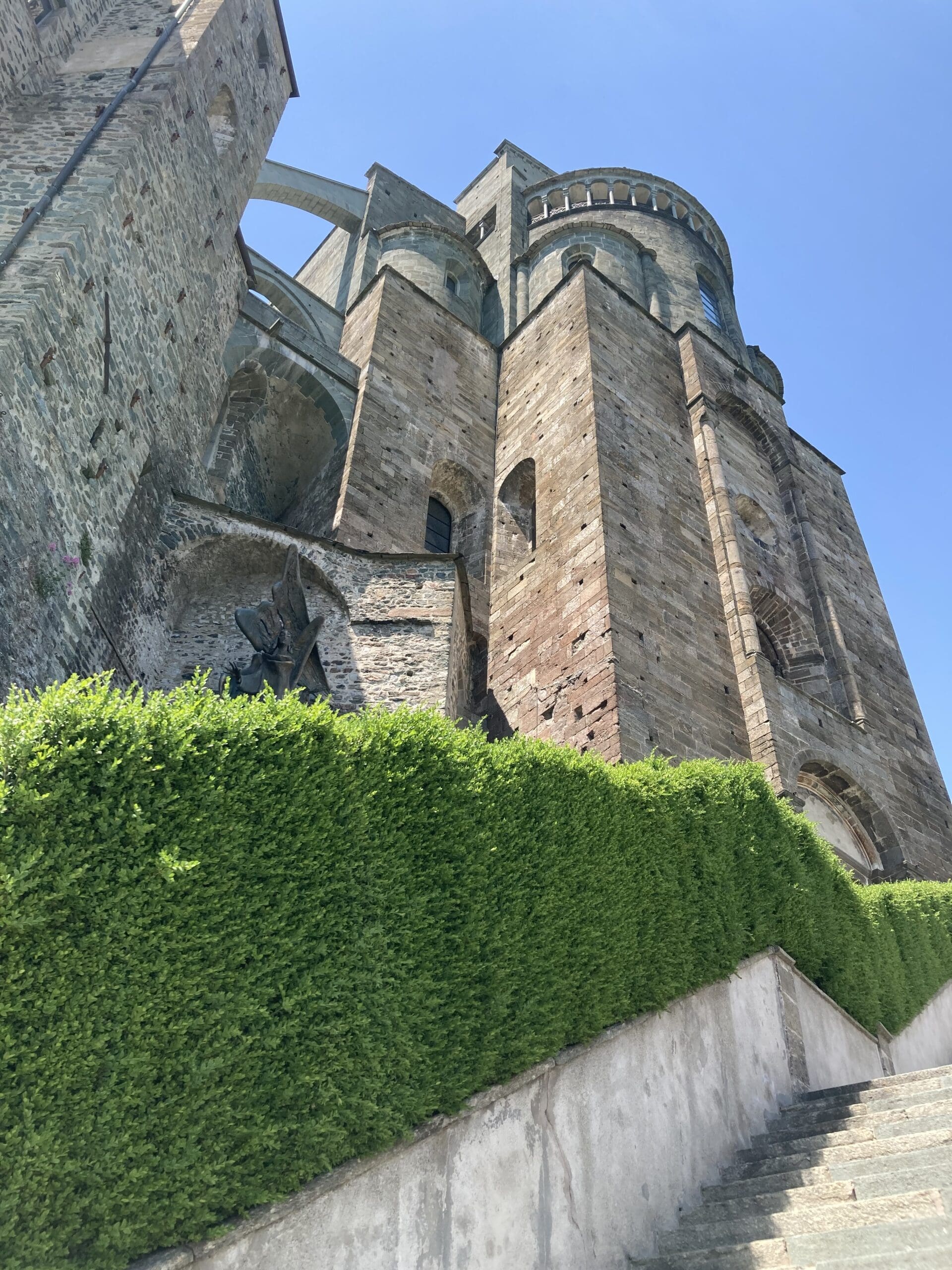 A tall stone building with arched windows and towers rises above a neatly trimmed green hedge and stone steps under a clear blue sky.