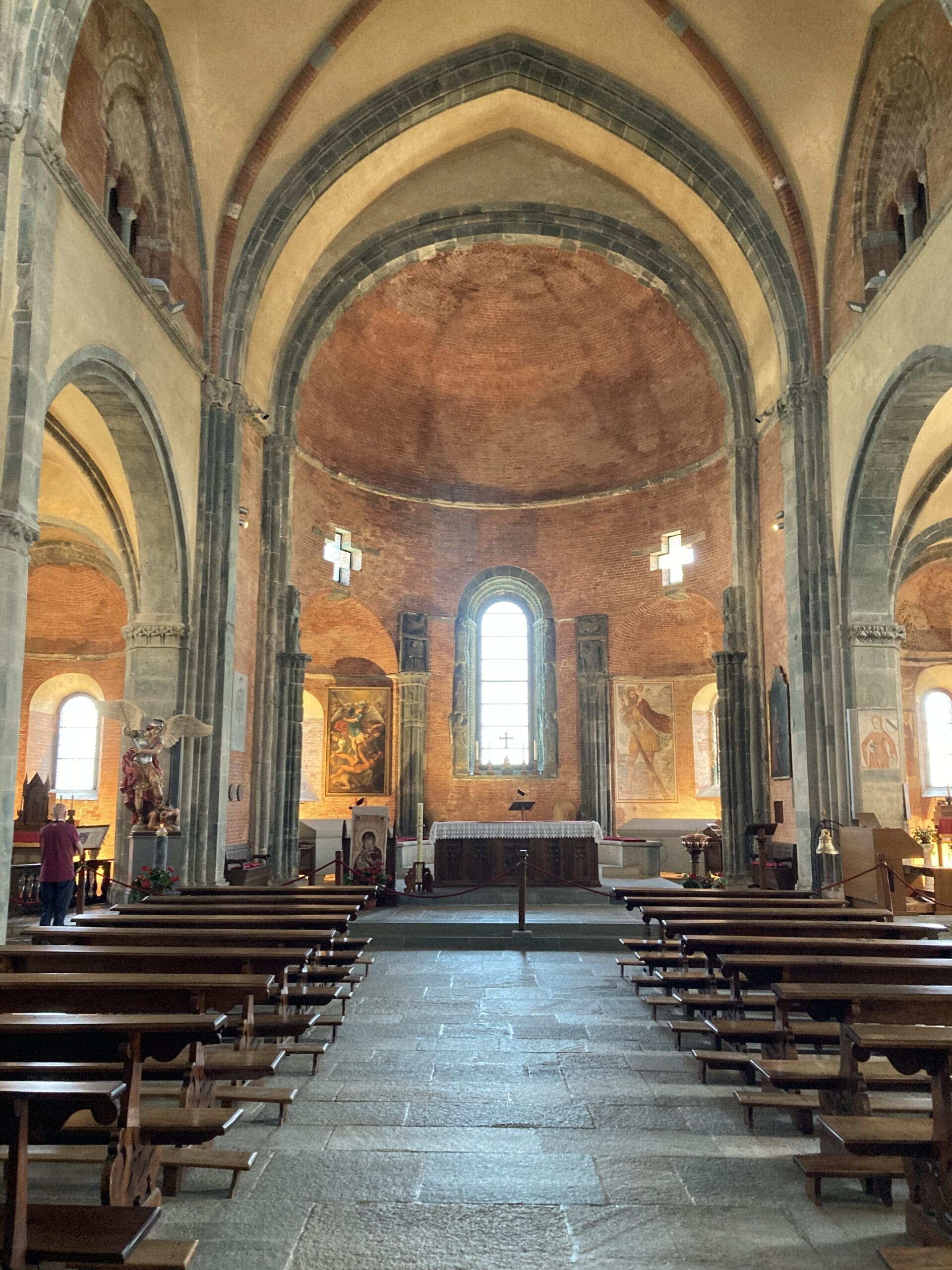 Interior of an old church with stone arches, wooden pews, paintings on the walls, and a central altar beneath a large arched window that lets in natural light.