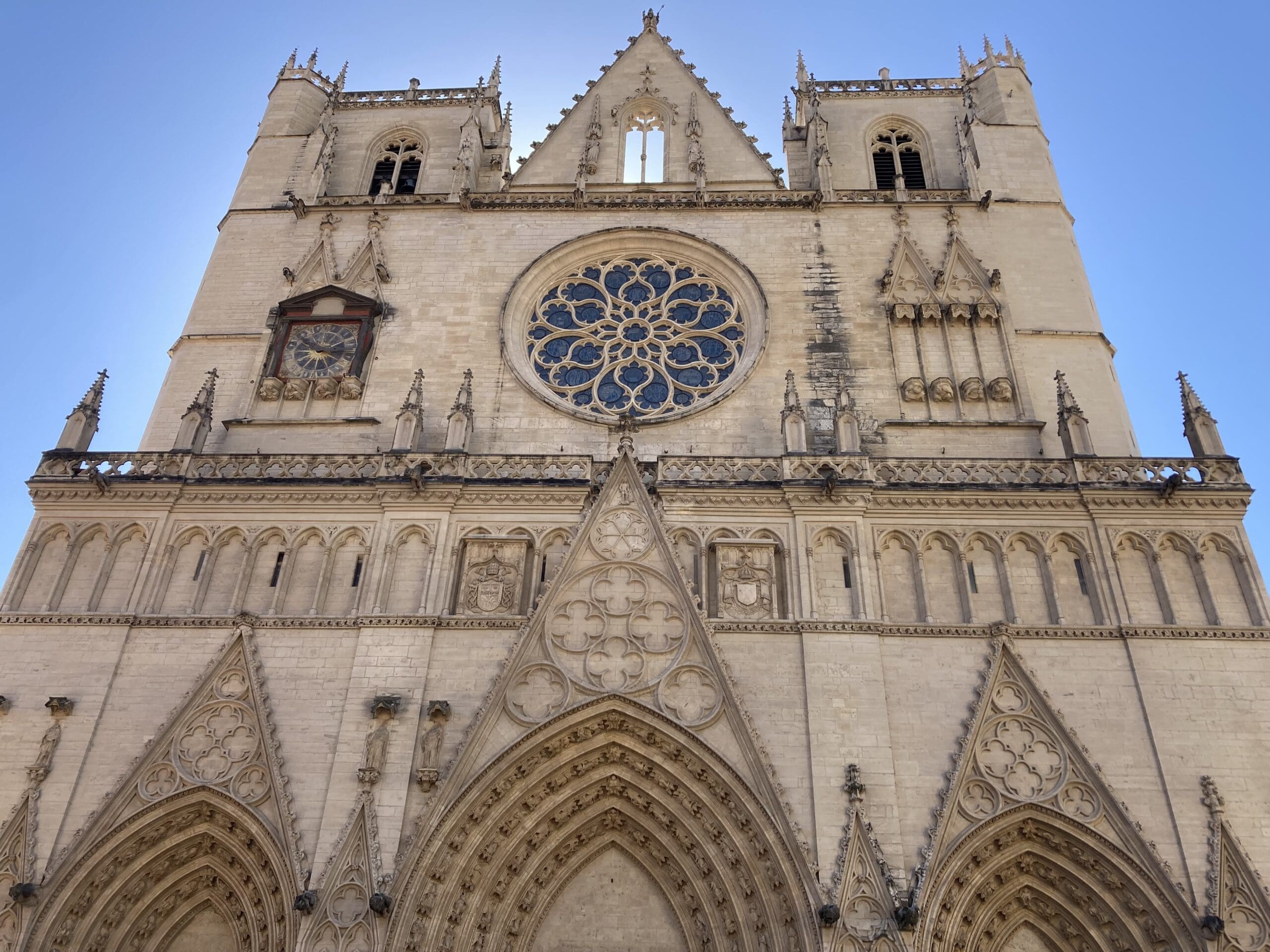 Facade of a Gothic cathedral with pointed arches, intricate stone carvings, and a large circular stained glass rose window. Two towers rise on either side under a clear blue sky.