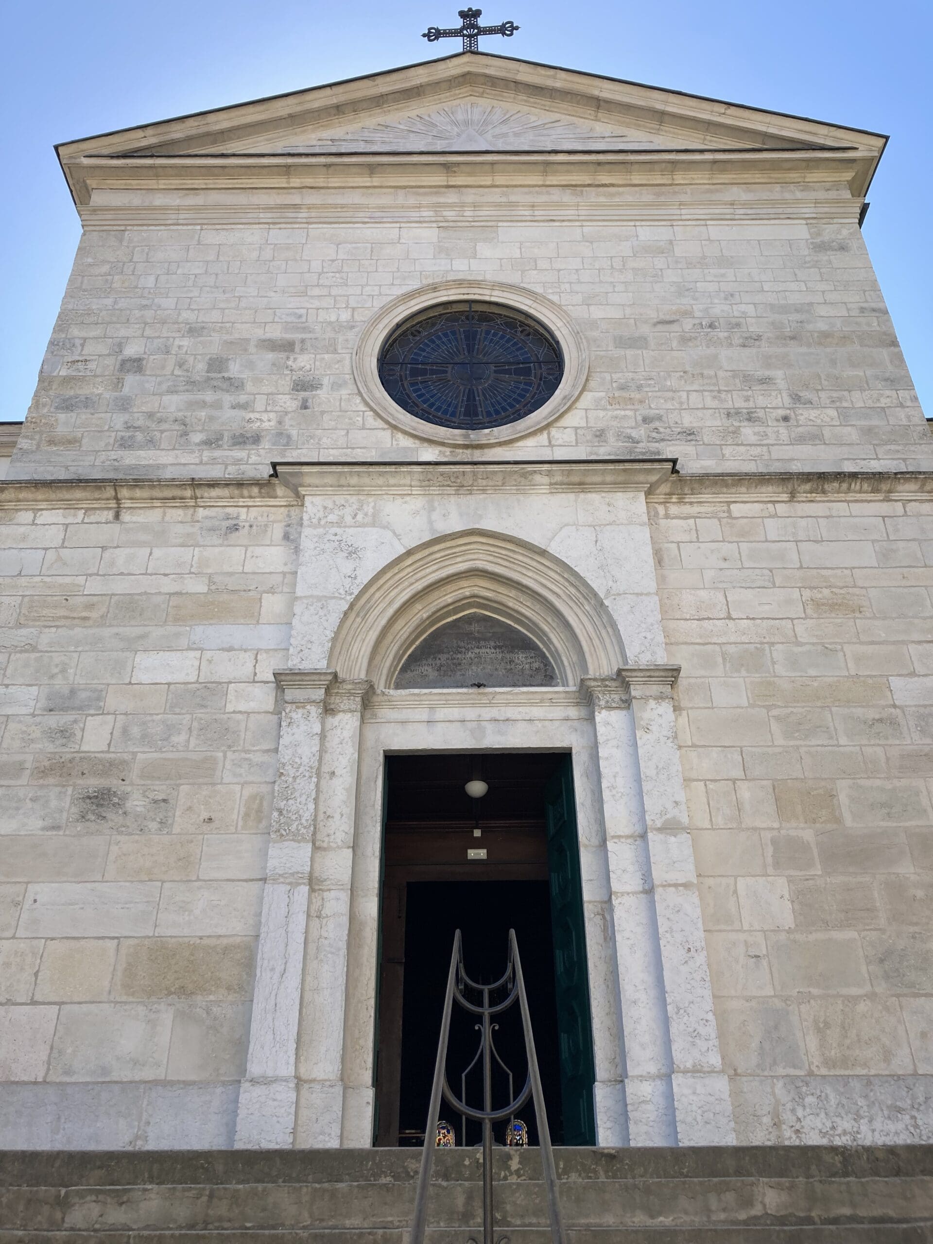 A stone church façade with an arched doorway, open green door, round stained glass window above, and a cross at the roof peak, seen from a low angle on a bright day.