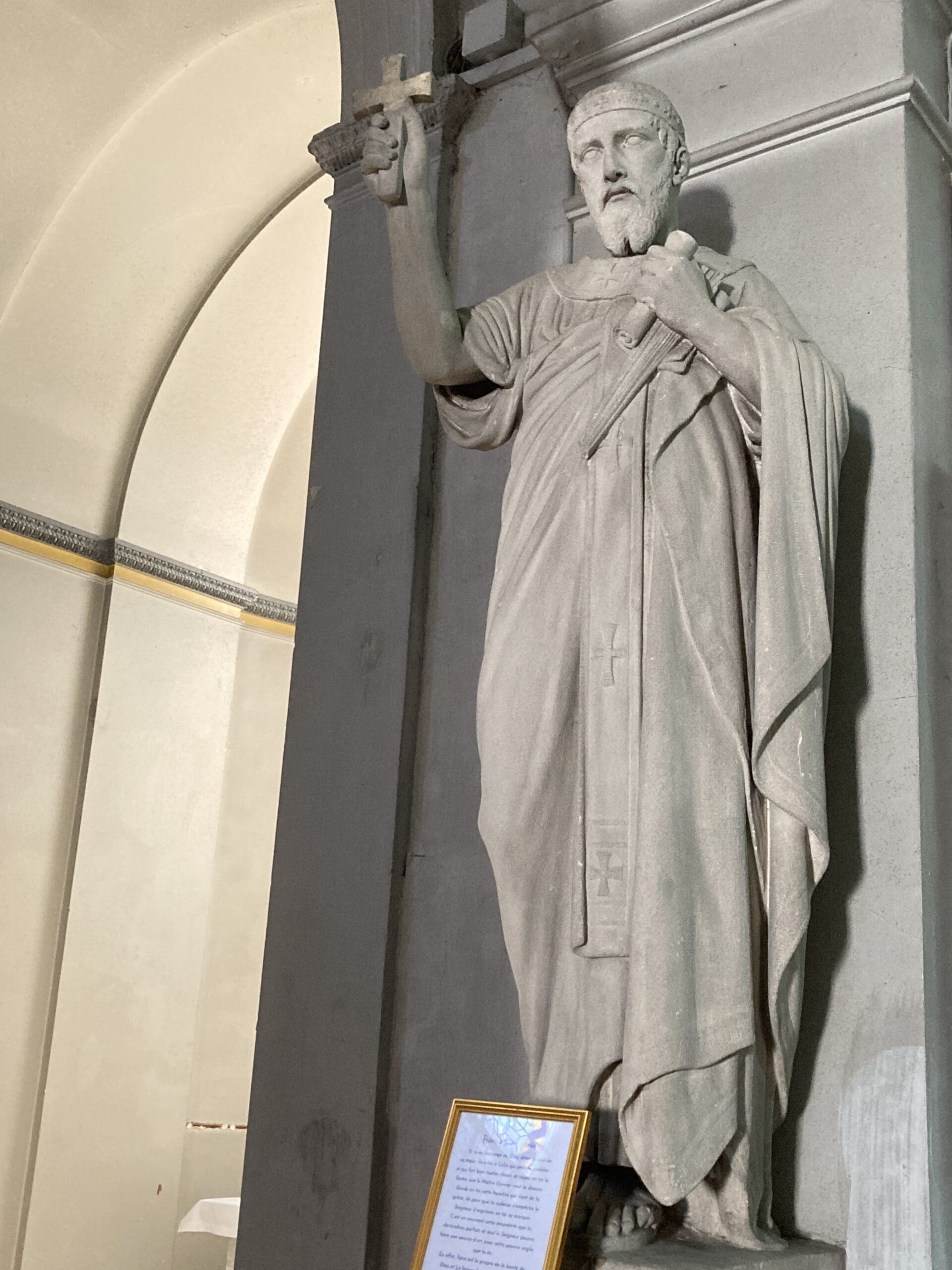 A stone statue of a robed bearded man holding a cross in his right hand. The statue stands indoors next to an arched wall, with a framed plaque placed at its base.