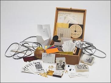 A wooden box filled with assorted items, including cards, papers, a pear, matches, a film reel, colored squares, and various small objects, all arranged on a white background.