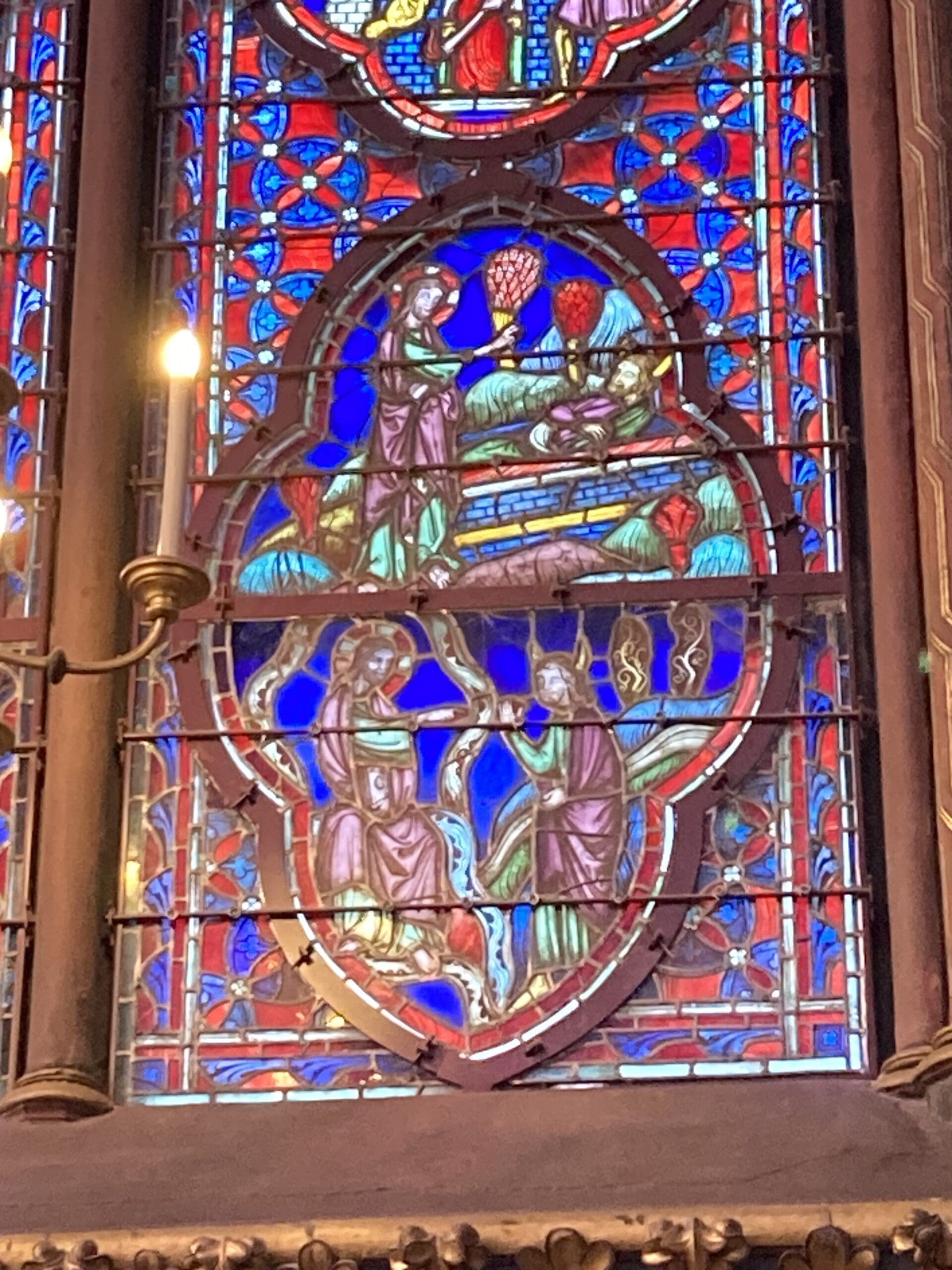 Stained glass window depicting medieval figures in colorful robes, with intricate red, blue, and green designs, divided into two main scenes, bordered by ornate tracery. A vertical lamp is visible on the left side.