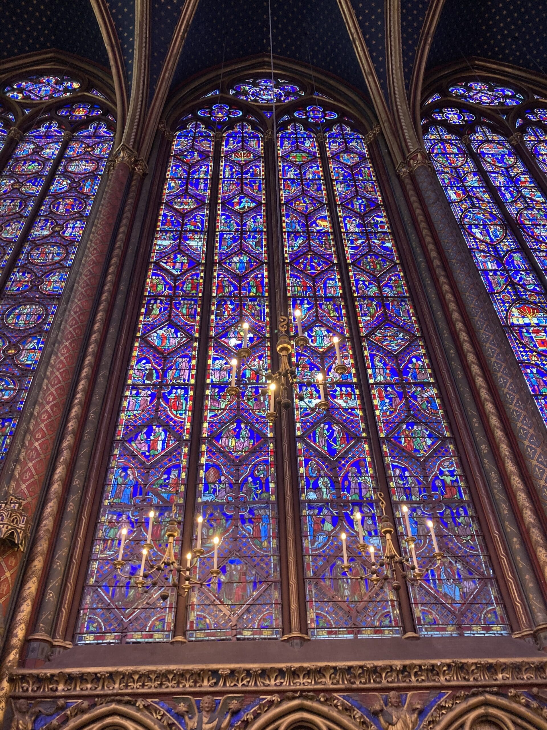 Tall stained-glass windows with intricate, colorful geometric and religious patterns, framed by ornate Gothic arches. A golden chandelier with lit candles hangs in front of the vibrant glass.