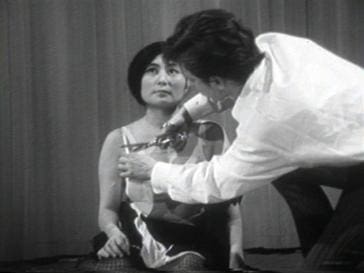 A person sits onstage looking up while another person uses scissors to cut pieces from their clothing. The scene appears to be from a performance or art event, with a curtain in the background.