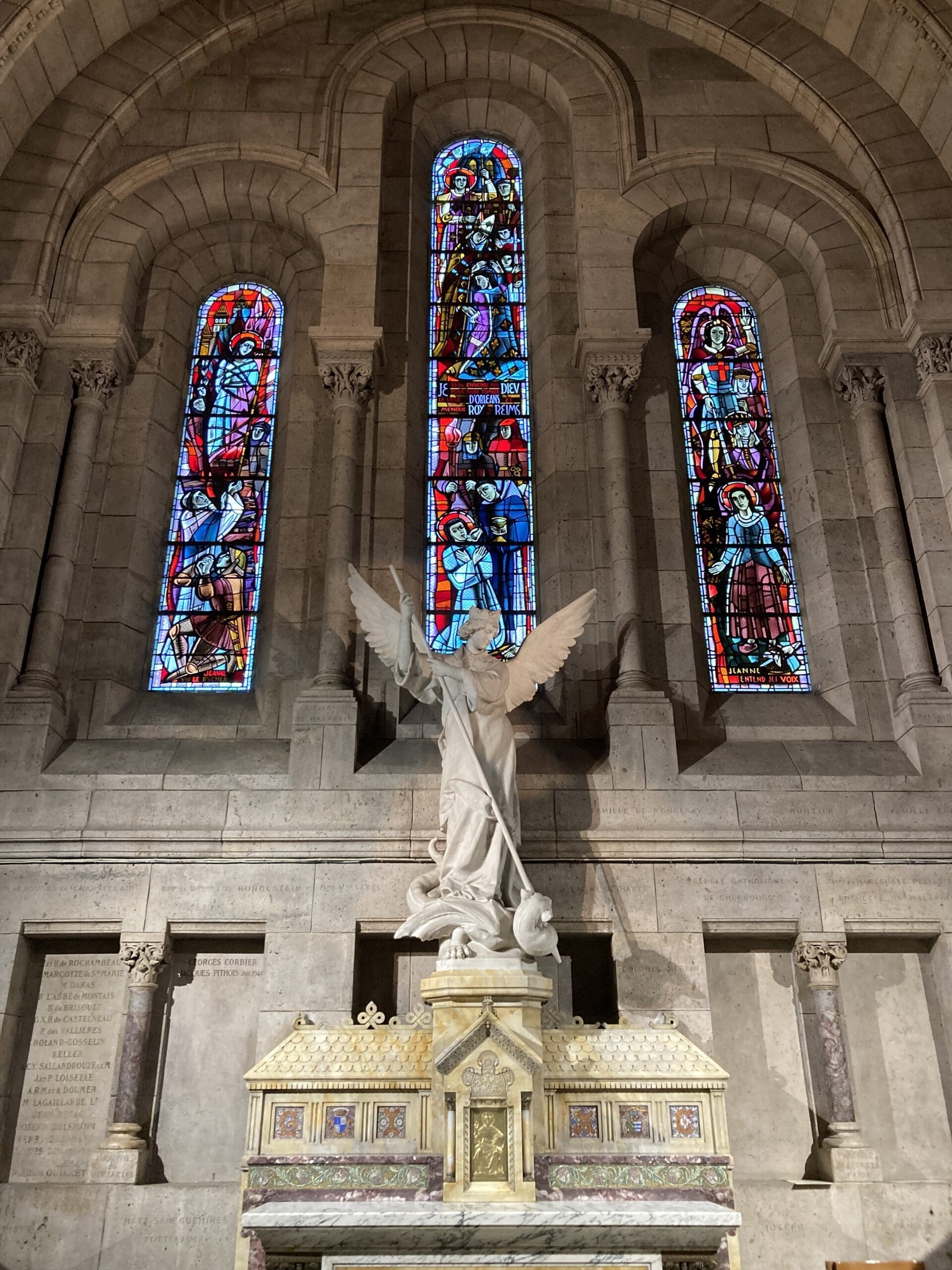 Stone angel statue with raised sword stands before three colorful stained glass windows inside a stone church. The altar below features ornate gold and marble decorations.