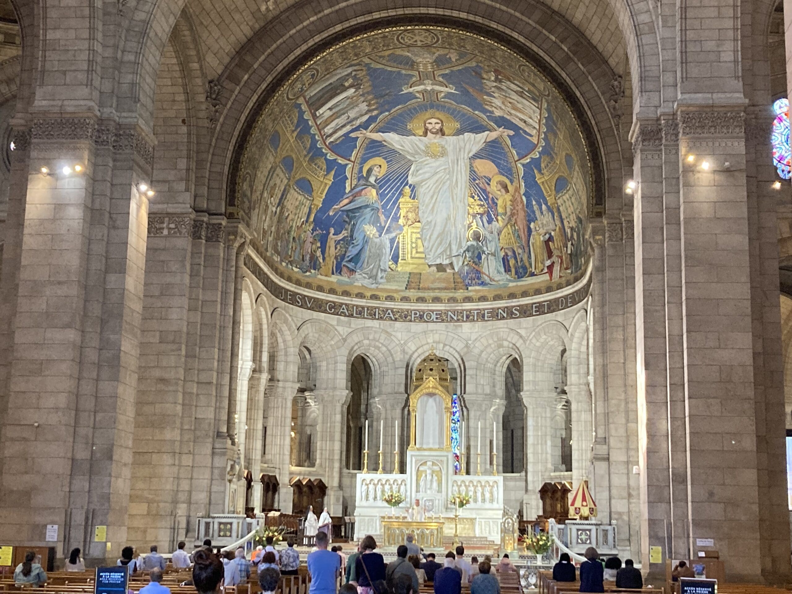 People sit in pews facing an ornate church altar with a large, colorful mosaic of Jesus and angels above. The stone interior is grand, with arches, columns, and glowing lights illuminating the artwork.