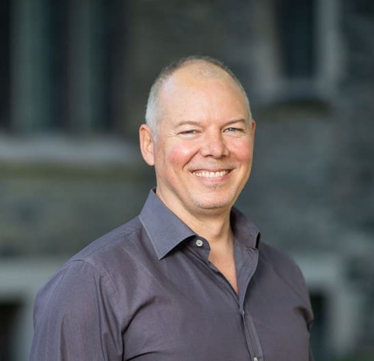 A smiling man with short light hair wearing a dark purple button-up shirt stands outdoors in front of a blurred stone building.