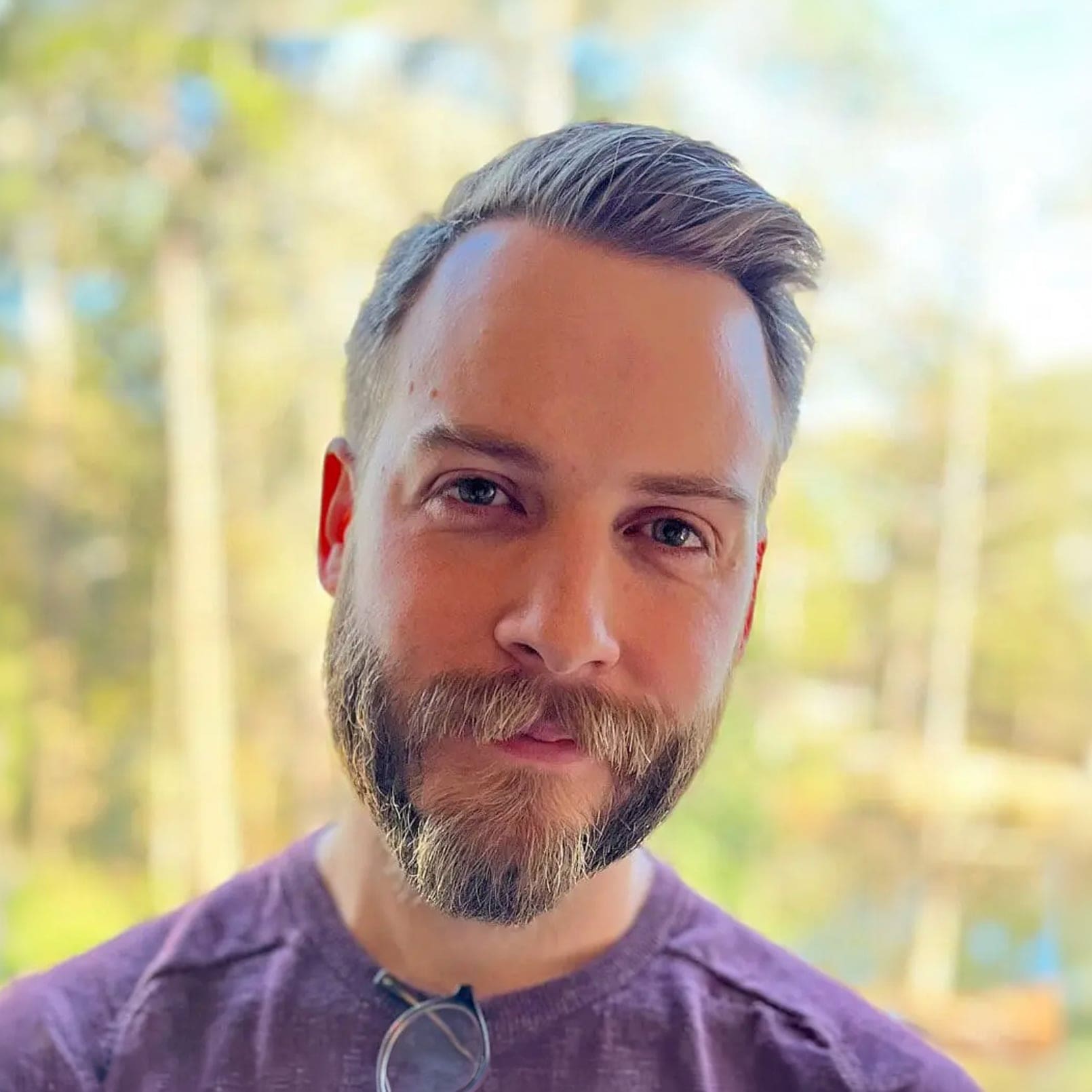 A man with light brown hair and a trimmed beard, wearing a purple shirt, stands outdoors in soft sunlight with a blurred background of trees and greenery.