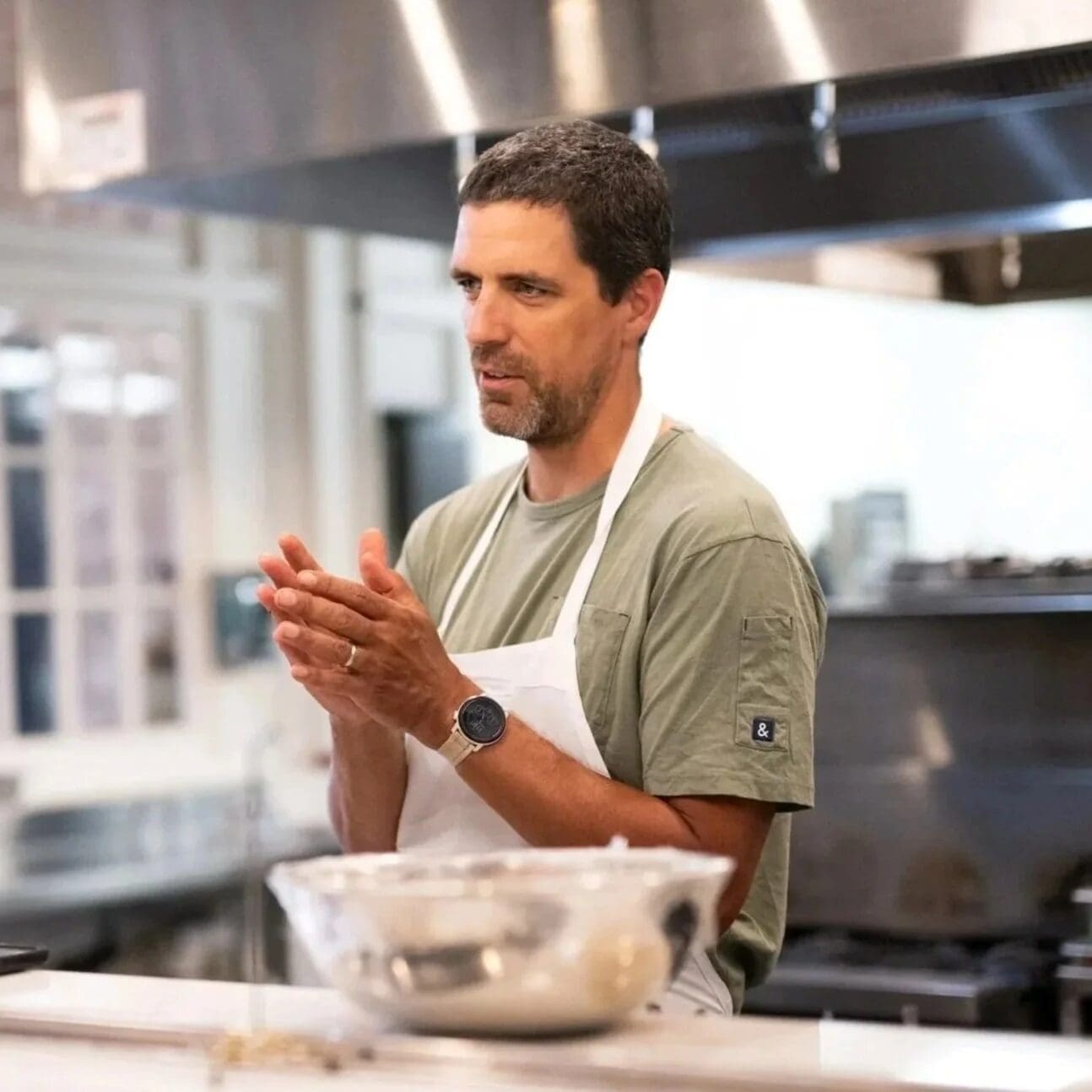 A man wearing a gray T-shirt and white apron stands in a kitchen, clapping his hands. A glass mixing bowl is on the counter in front of him, and stainless steel kitchen equipment is in the background.